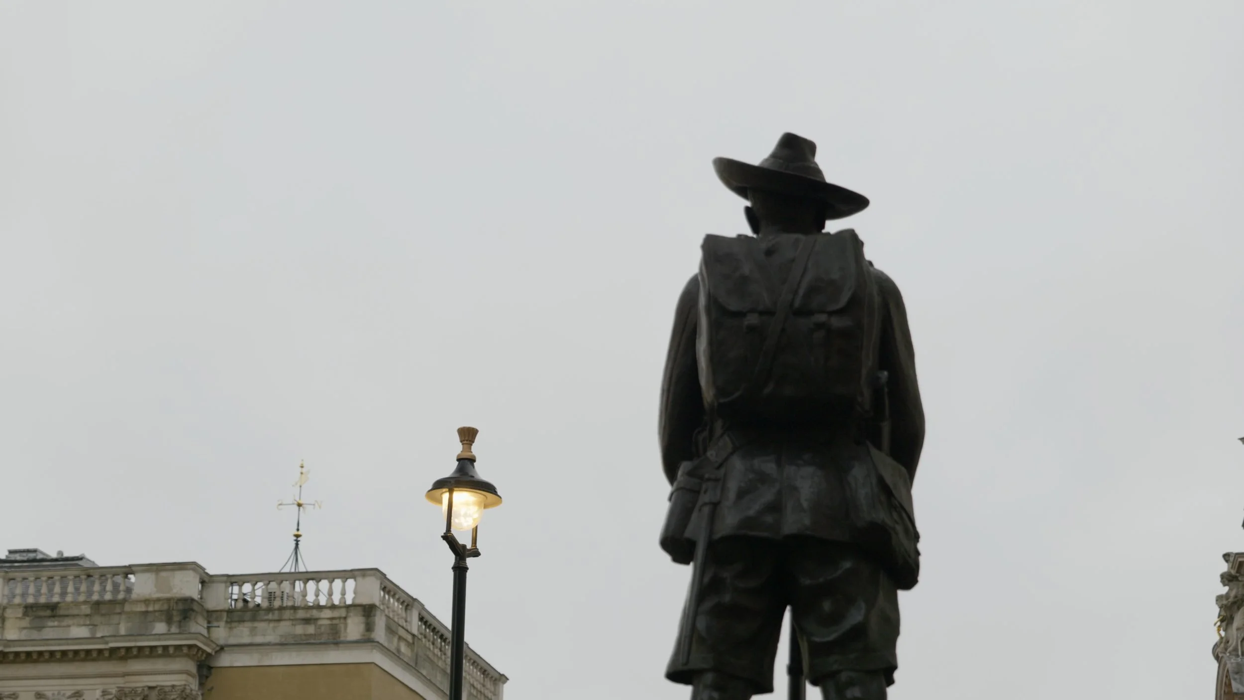 Statue of a man wearing a hat and a backpack, seen from behind, in front of historic buildings with a streetlamp, overcast sky.