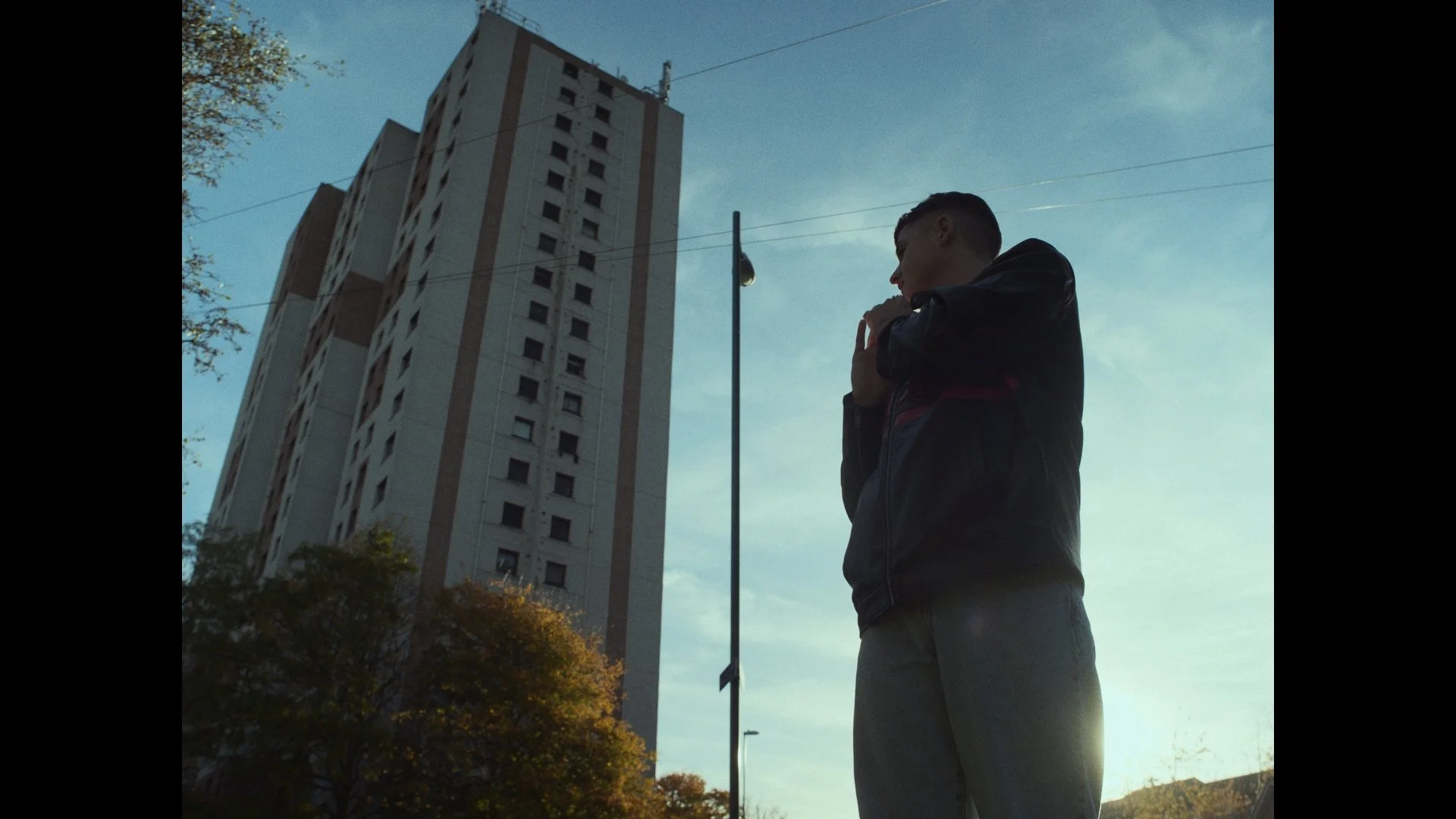 A young man standing outdoors at sunset, wearing a black jacket and gray pants, with a tall building and a streetlight in the background.