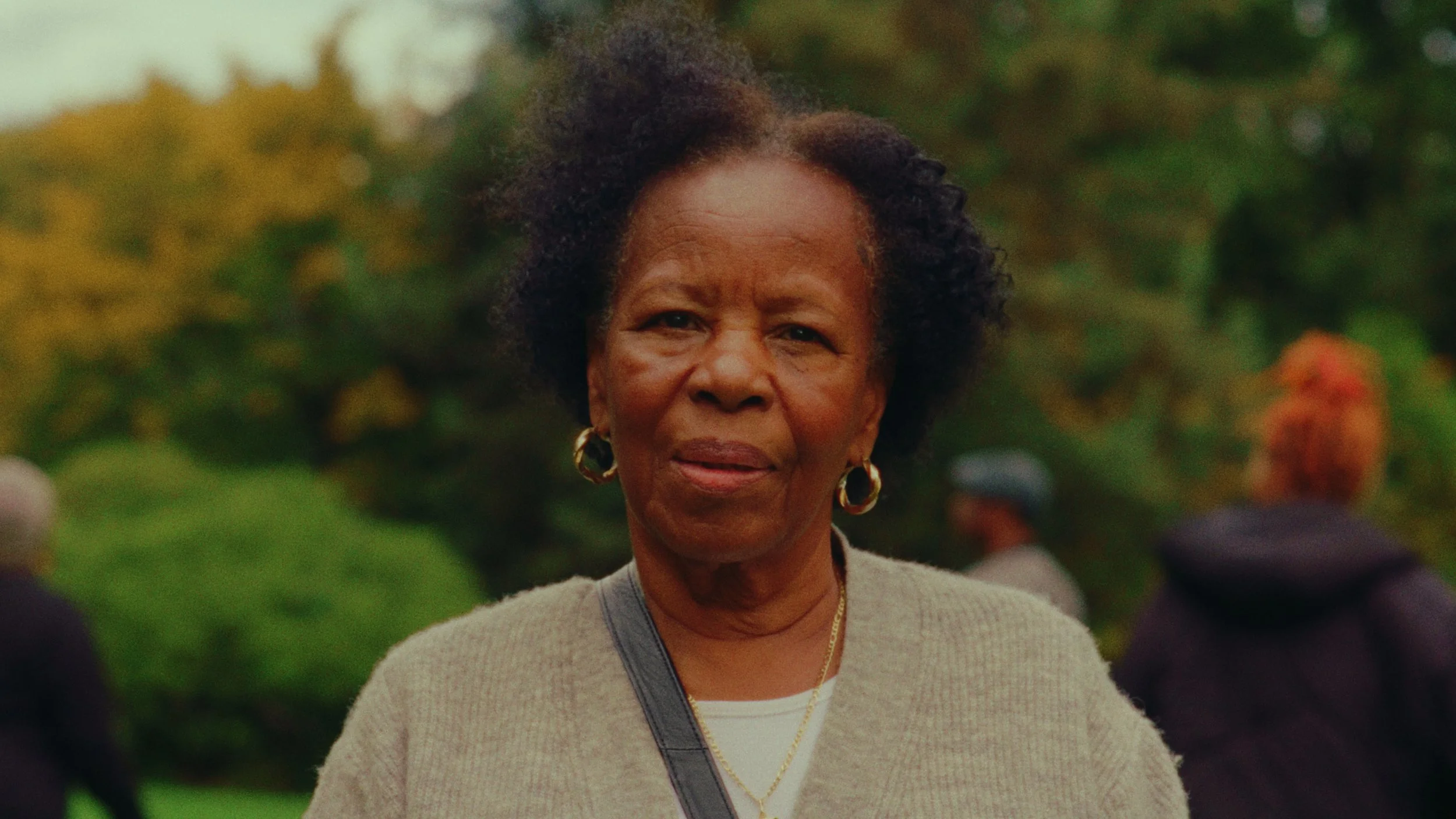 Close-up of an elderly woman with short curly hair, wearing gold hoop earrings, a beige sweater, and a gold necklace, outdoors with blurred greenery and other people in the background.