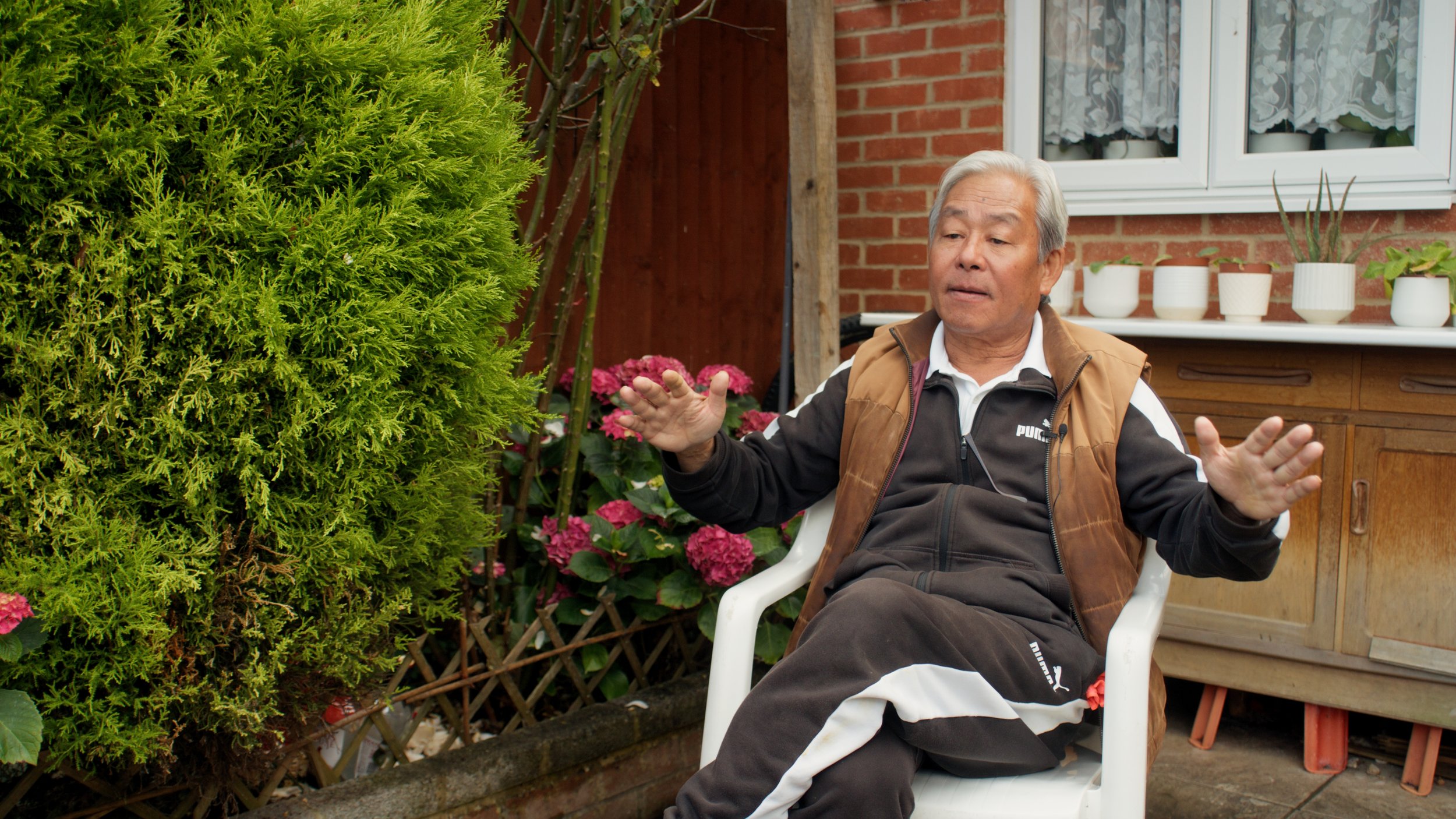 An elderly man sitting outdoors in a garden, gesturing with his hands while talking. There are pink flowers and green bushes beside him, and a brick house with white-framed windows in the background.