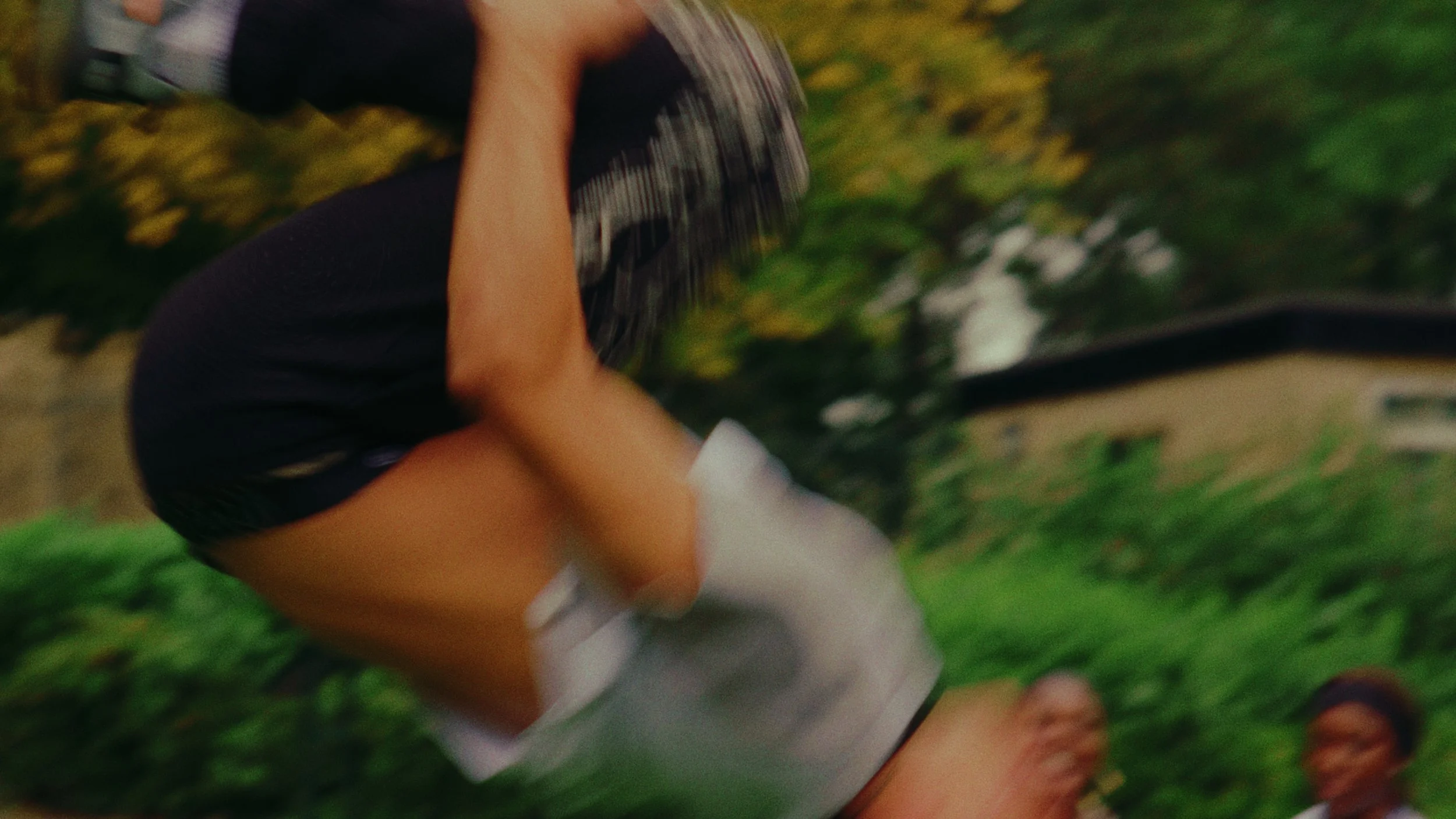 A person wearing black shorts and a white tank top holding a skateboard, with a house and greenery in the background.
