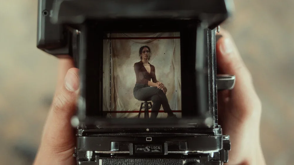 A woman sitting on a stool, being photographed through the viewfinder of a large professional camera.