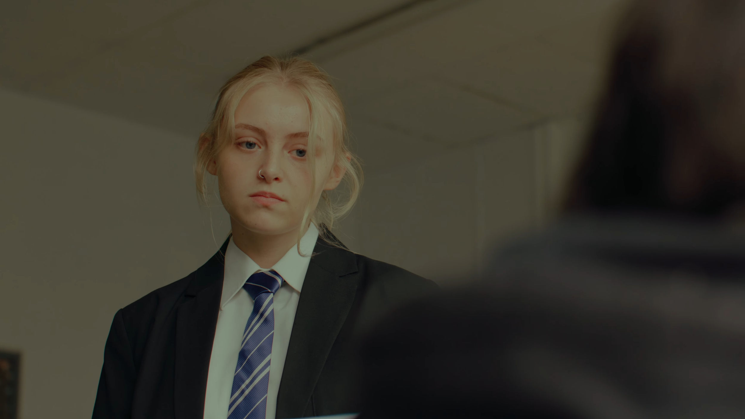 A young woman with blonde hair wearing a black blazer, white shirt, and striped tie looking down with a serious expression.
