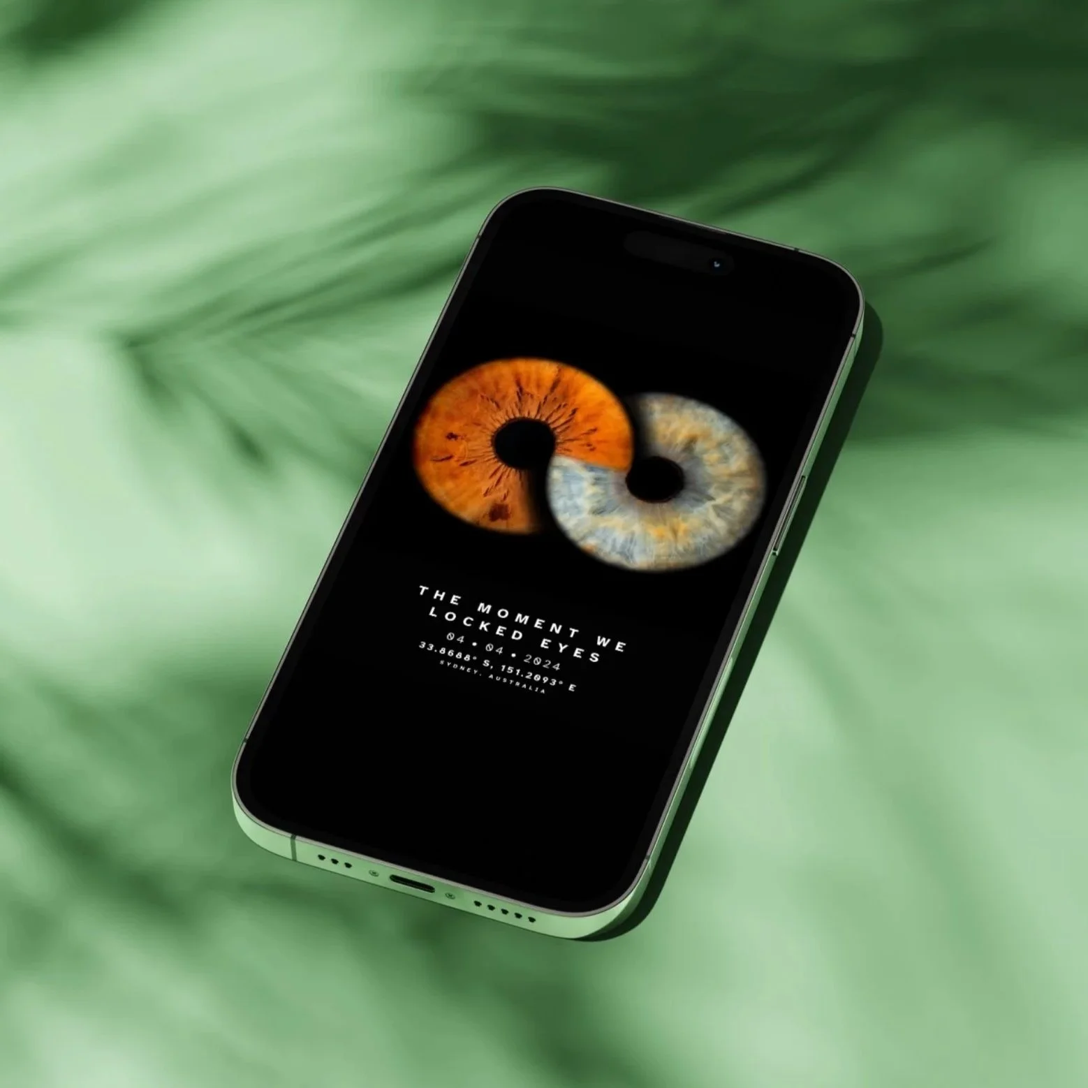 A smartphone displaying a lock screen with a high-resolution image of two human irises, one brown and one blue, and text that reads, "The moment we locked eyes," along with a date, time, and geographic coordinates, resting on a blurred green leafy background.