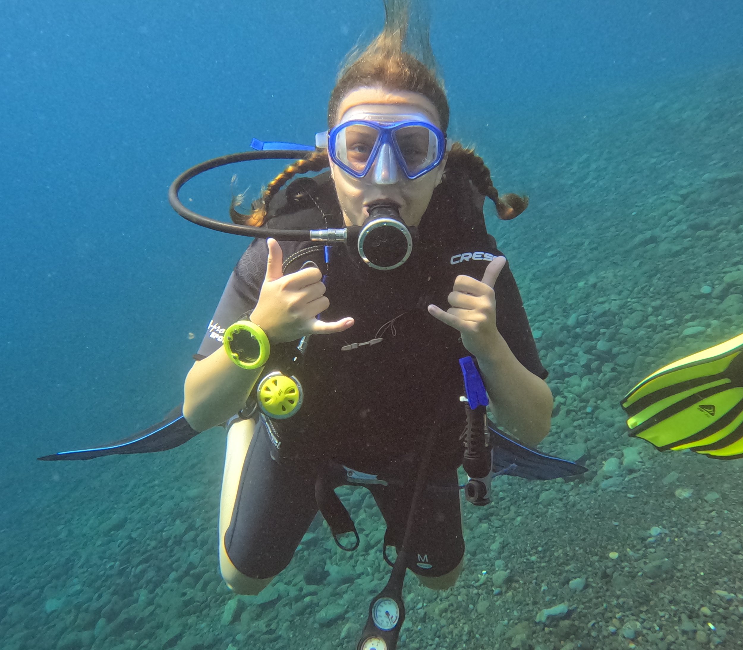 En person dykker under vand med scuba-udstyr og laver håndtegn. Personen har blå dykkermaske, våddragt og bruger dykkerudstyr.