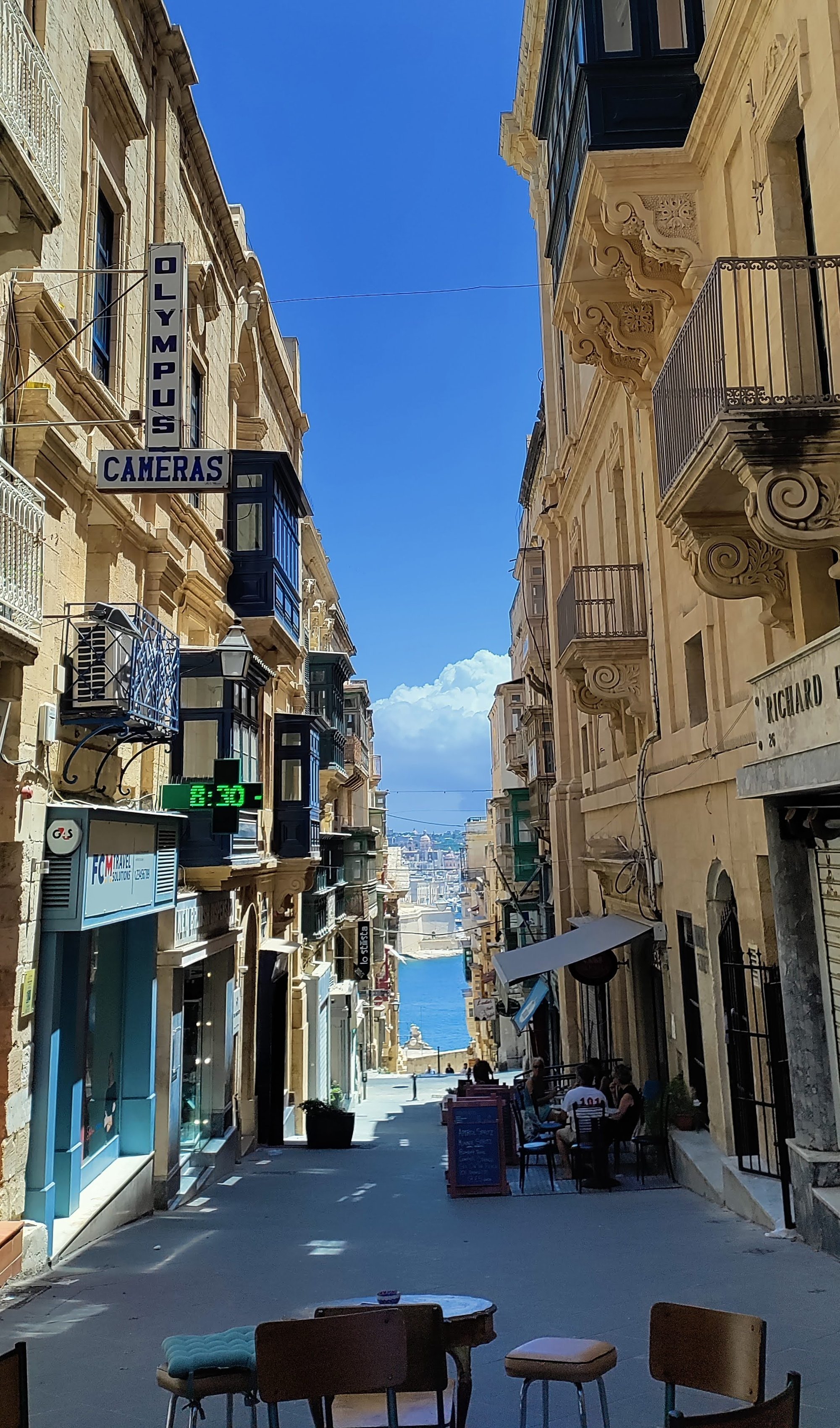 En smal gata kantad av gamla byggnader, kaféer med uteservering och utsikt över hamnen i fjärran under en klarblå himmel.