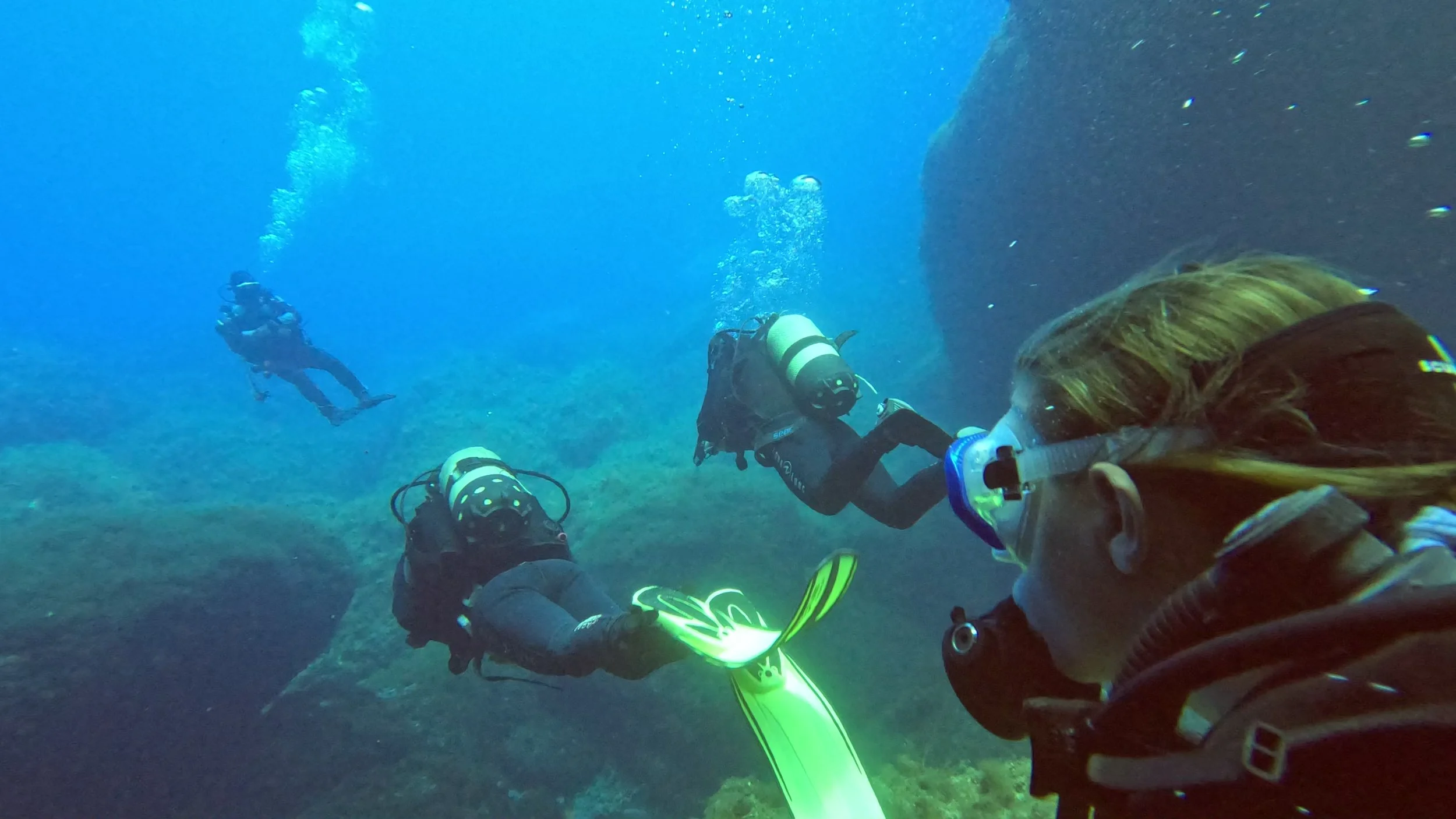 tre dykkere svømmer mod deres instruktør der ligger længere fremme i vandet og venter på dem over dem er det blå hav og under dem massive klippeformationer