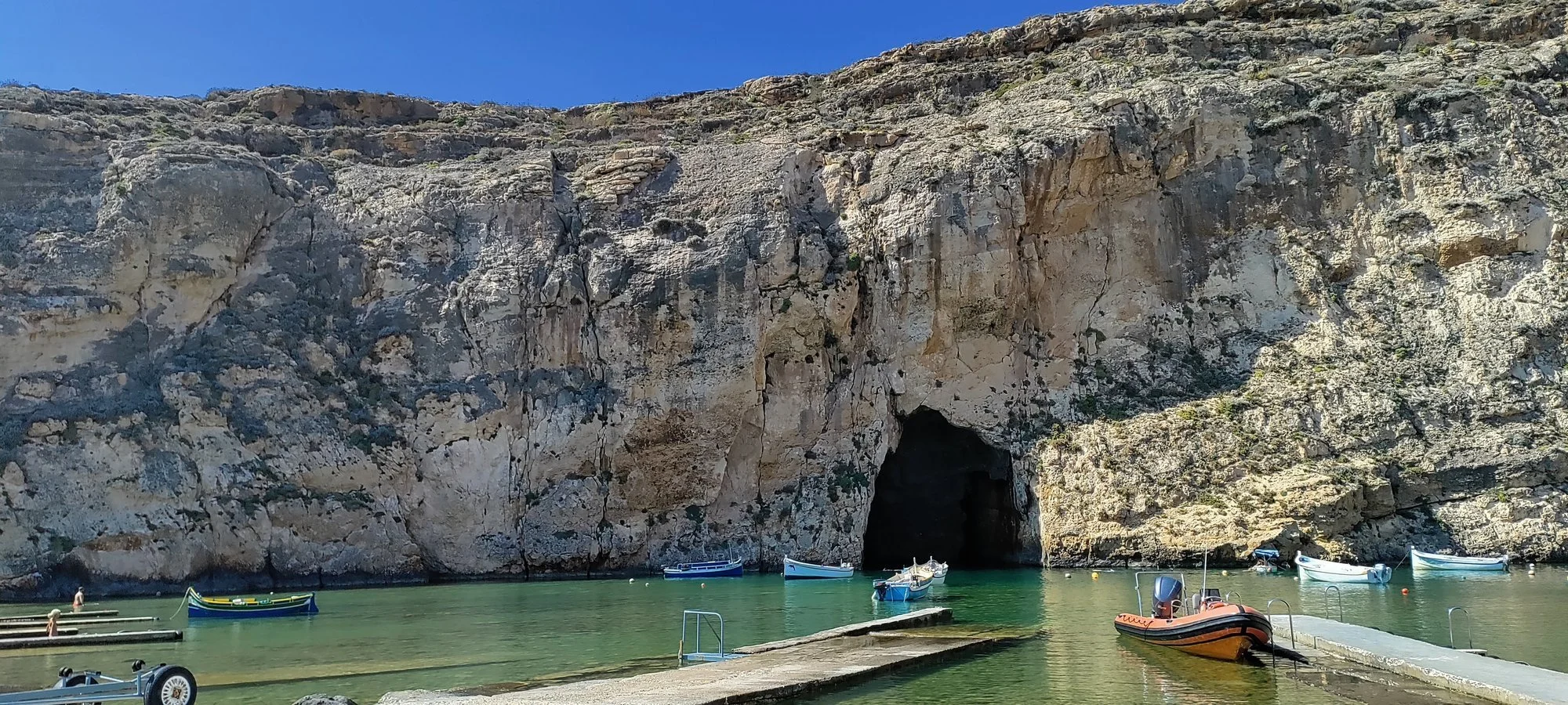 Lugnt inlandshav med klippformationer och grottor, flera små båtar och en motorbåt vid en brygga. Solen skiner från en klar himmel.