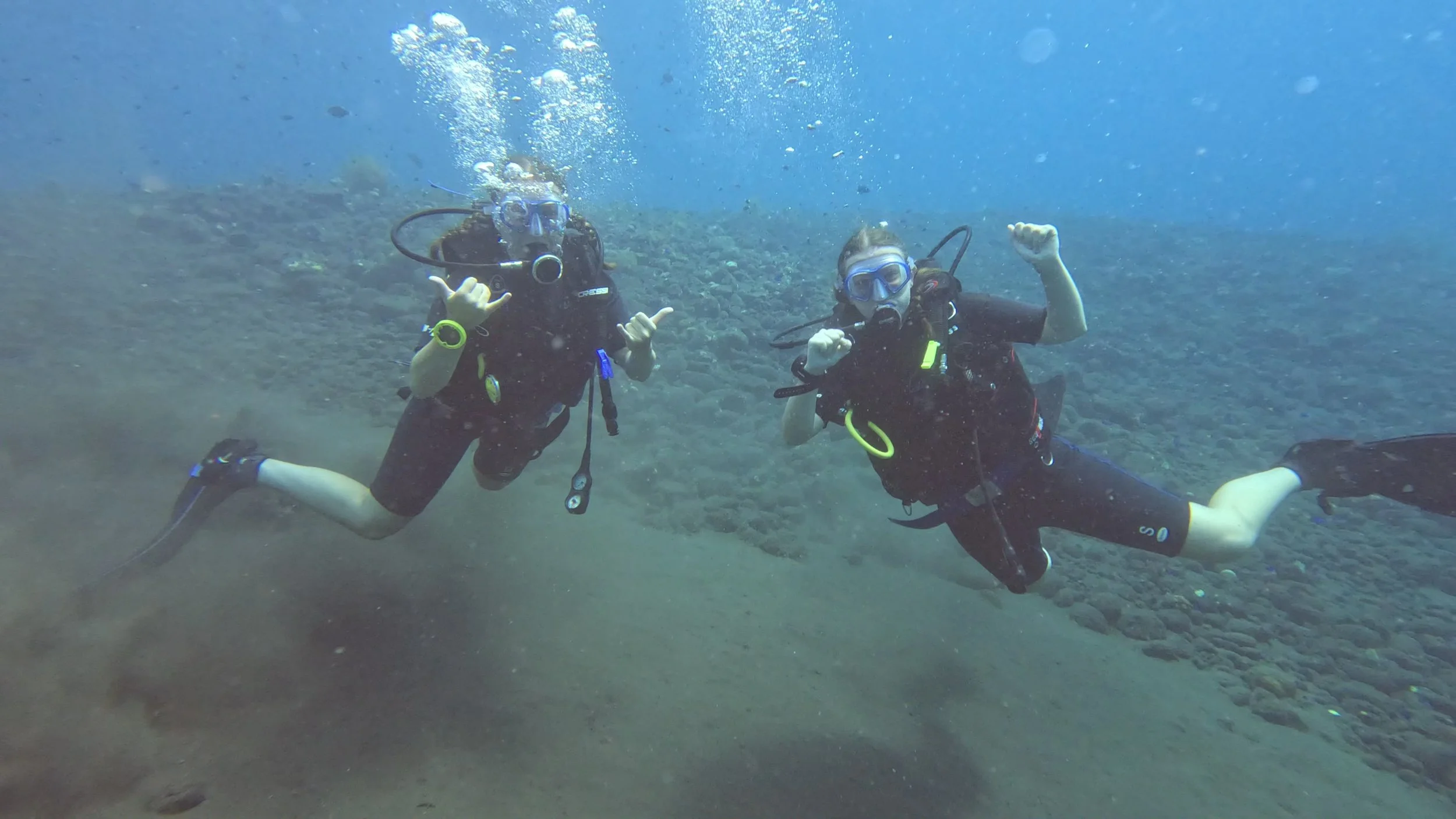 To dykkere under vandet på Bali giver håndsignaler og smiler i det klare blå hav – en oplevelse fyldt med eventyr og fælles glæde.