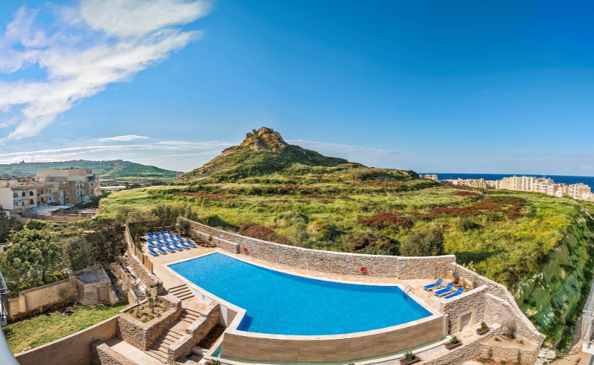 Pool med solstolar framför gröna bergskedjor med en stad i bakgrunden under en blå himmel.