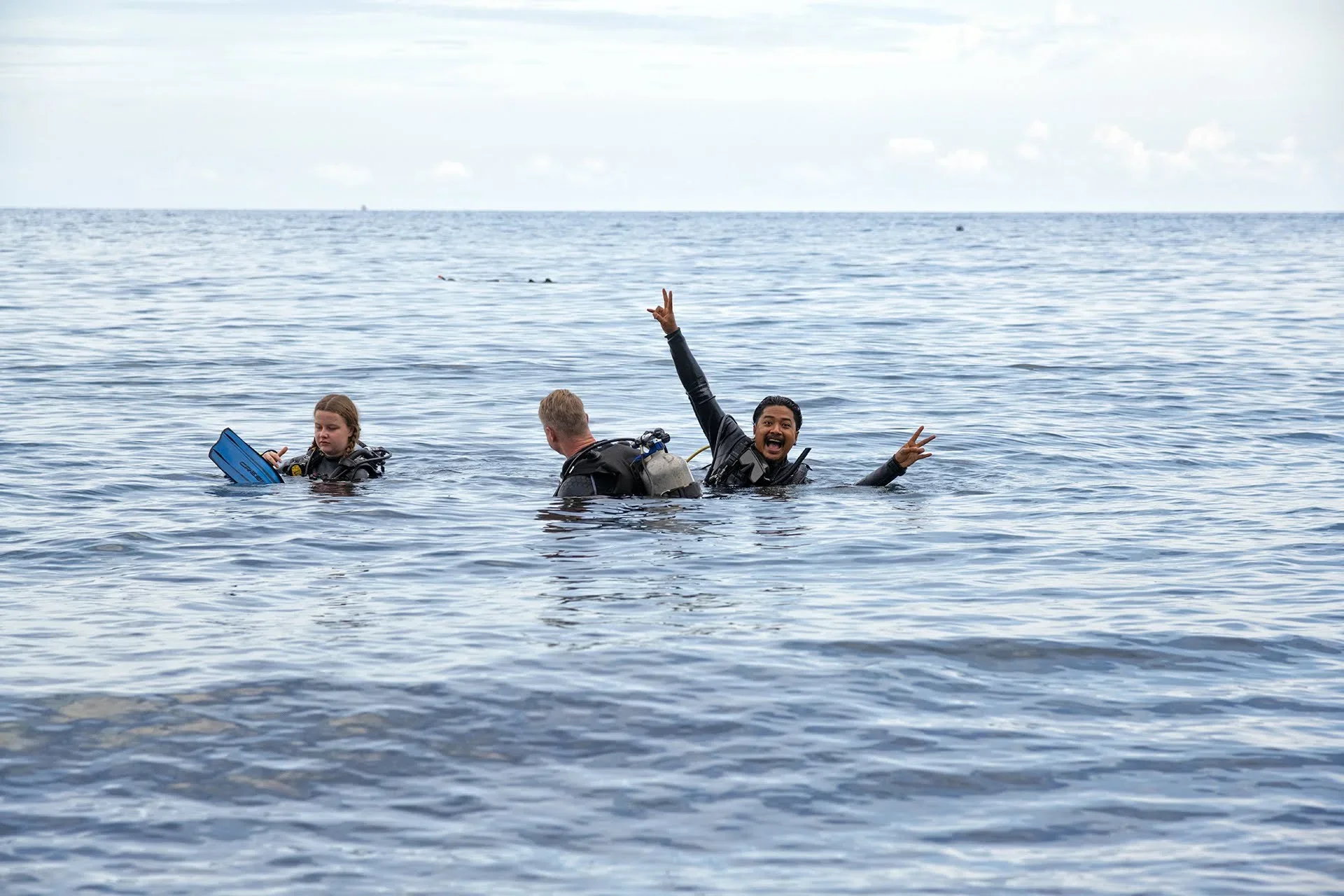 Tre personer i havet, to mænd og en pige, hvor den ene mand er glad og laver peace-tegn med hænderne.