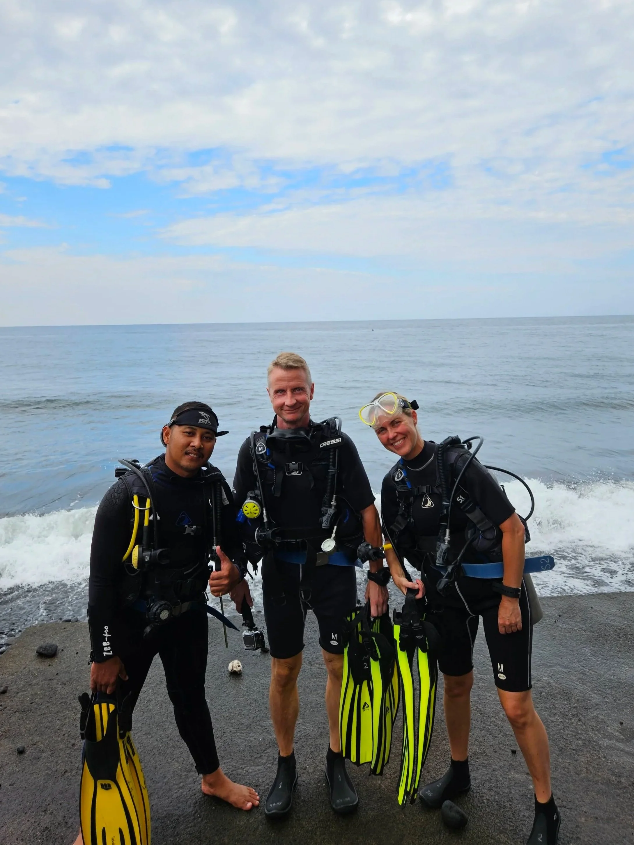 Tre dykkere står på en sort strand ved havet, iført dykkerudstyr og holder skarpe snorkler, klar til dykning.