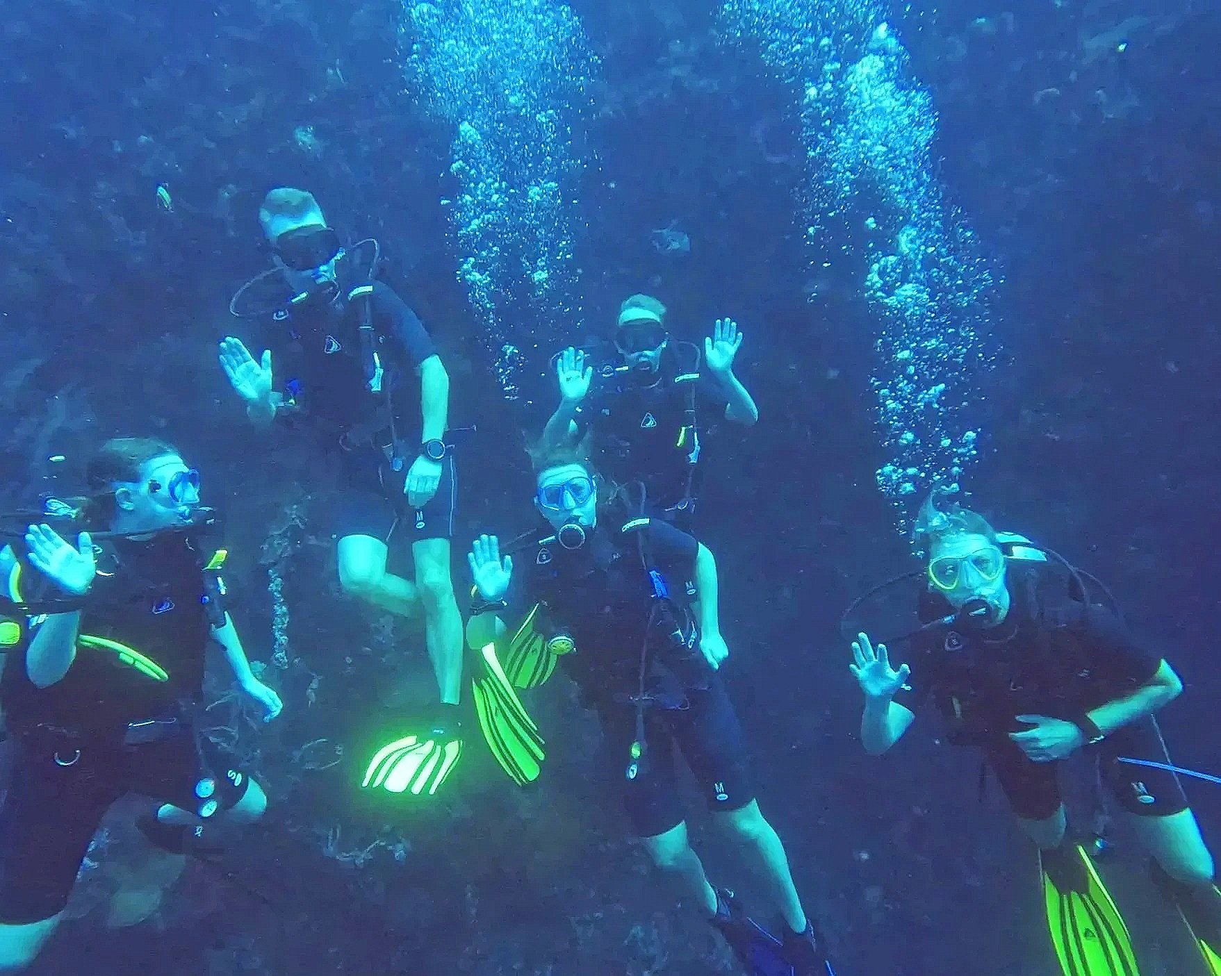 Gruppemedlemmer i dykkermaske og våddragt svømmer under vandet og hilser med hænderne