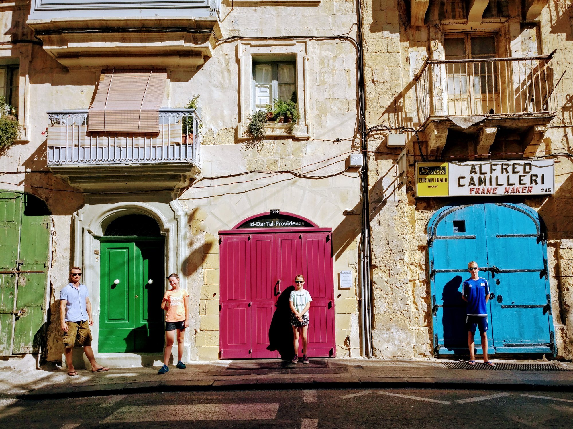 Fyra personer står framför färgade dörrar framför en gammal byggnad med fönster och balkonger. Det finns också en skylt med namnet "Alfred Camilleri" och en reklam för "Swish".