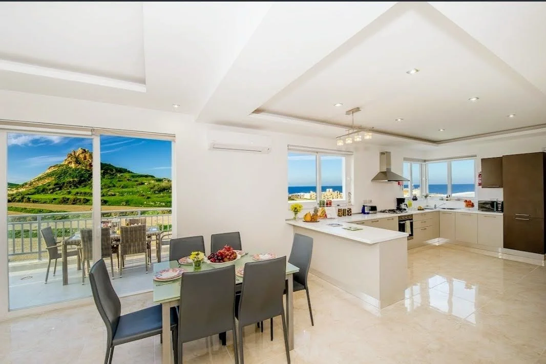 Moderne køkken og spisestue med udsigt til havet og grøn bjerglandskab gennem store vinduer og glasdør.