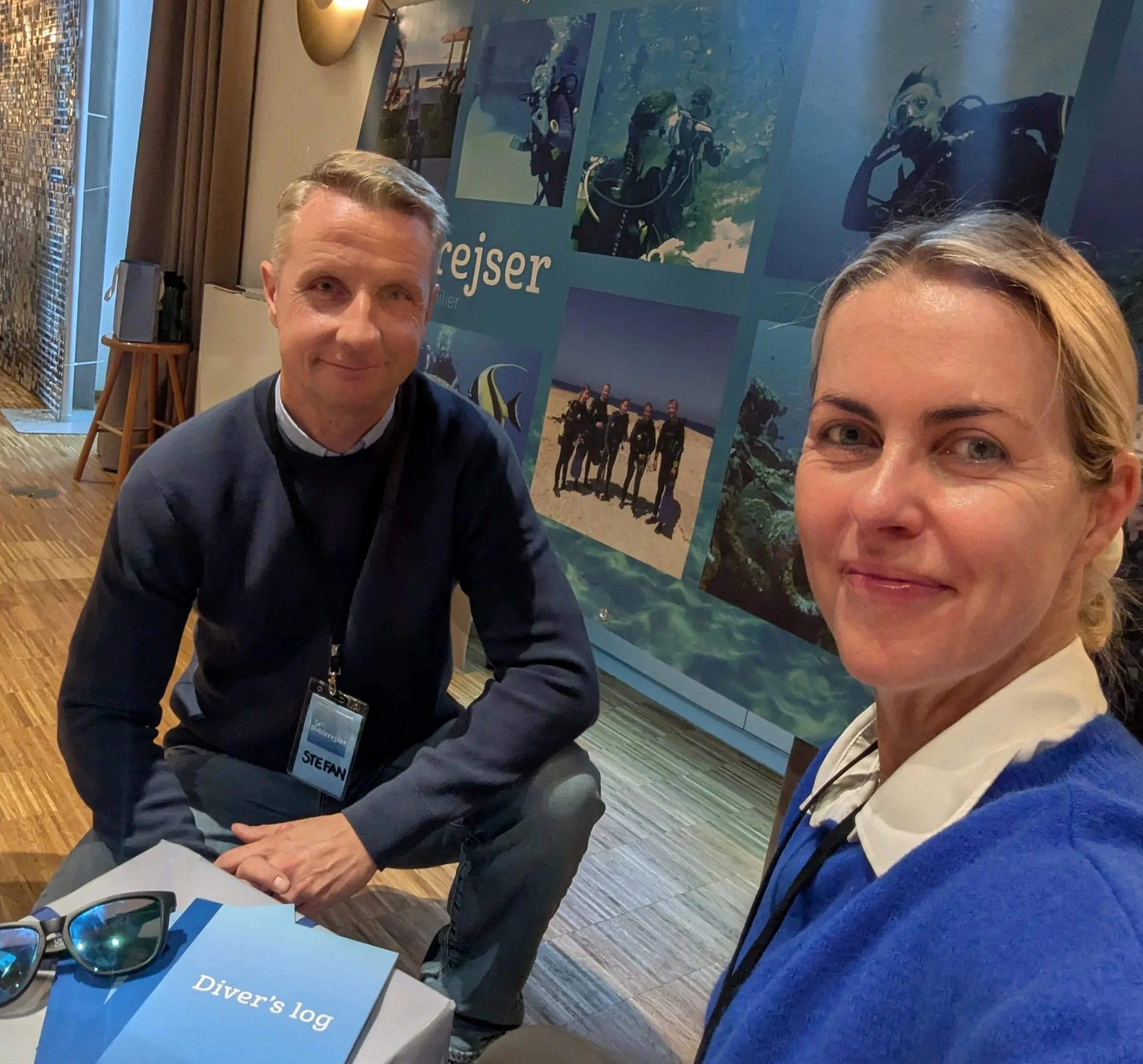 Två personer, en man och en kvinna, tar en selfie i ett rum med en utställningspanel bakom sig som visar bilder på dykning och resor.