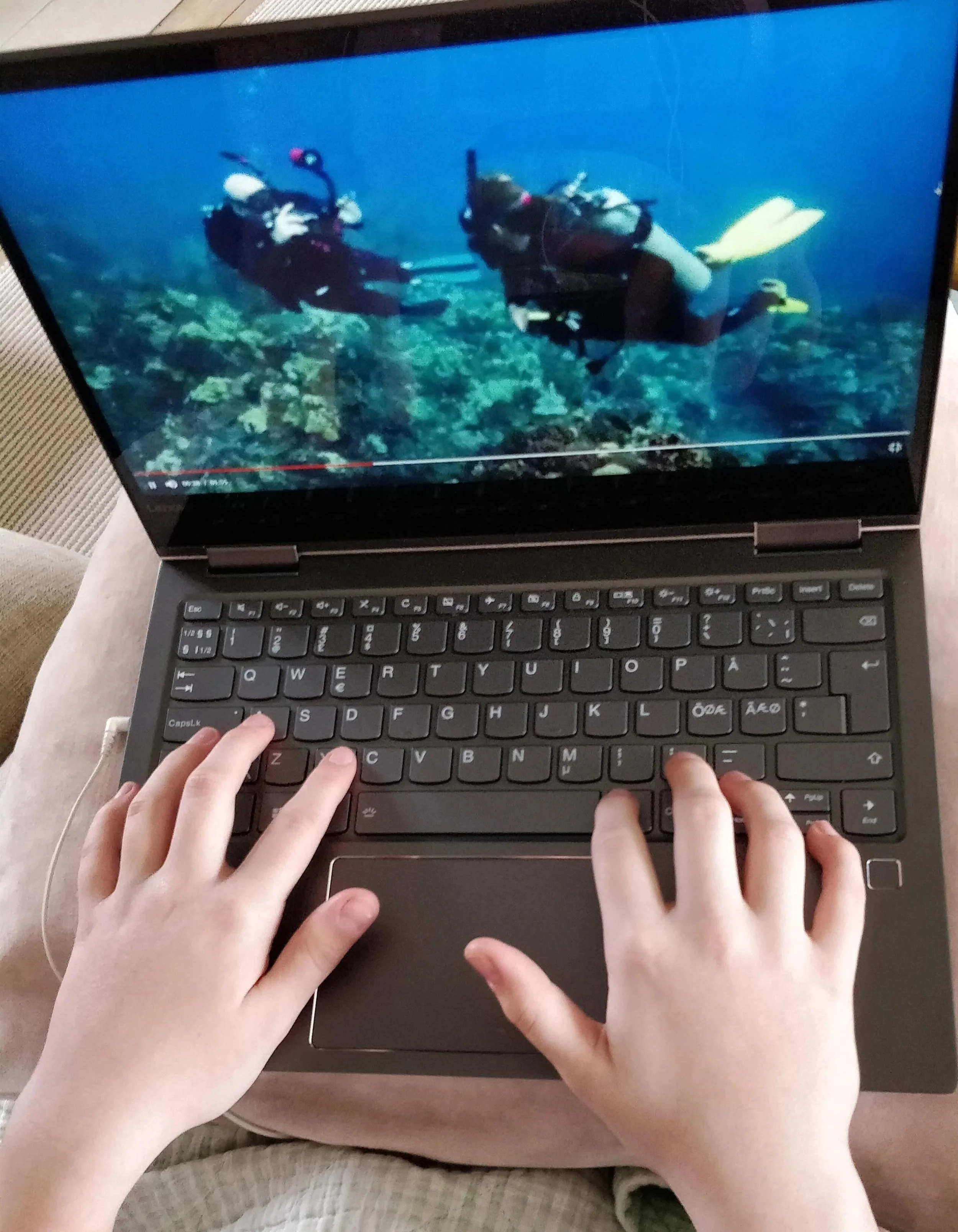 En ung persons hænder skriver på en bærbar computer, der viser en video med to dykkere under vandet - det er online undervisning til dykkercertifikat