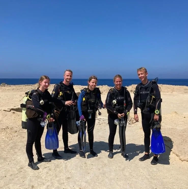 Fem personer klædt i dykkerudstyr står på en strand med havet i baggrunden.
