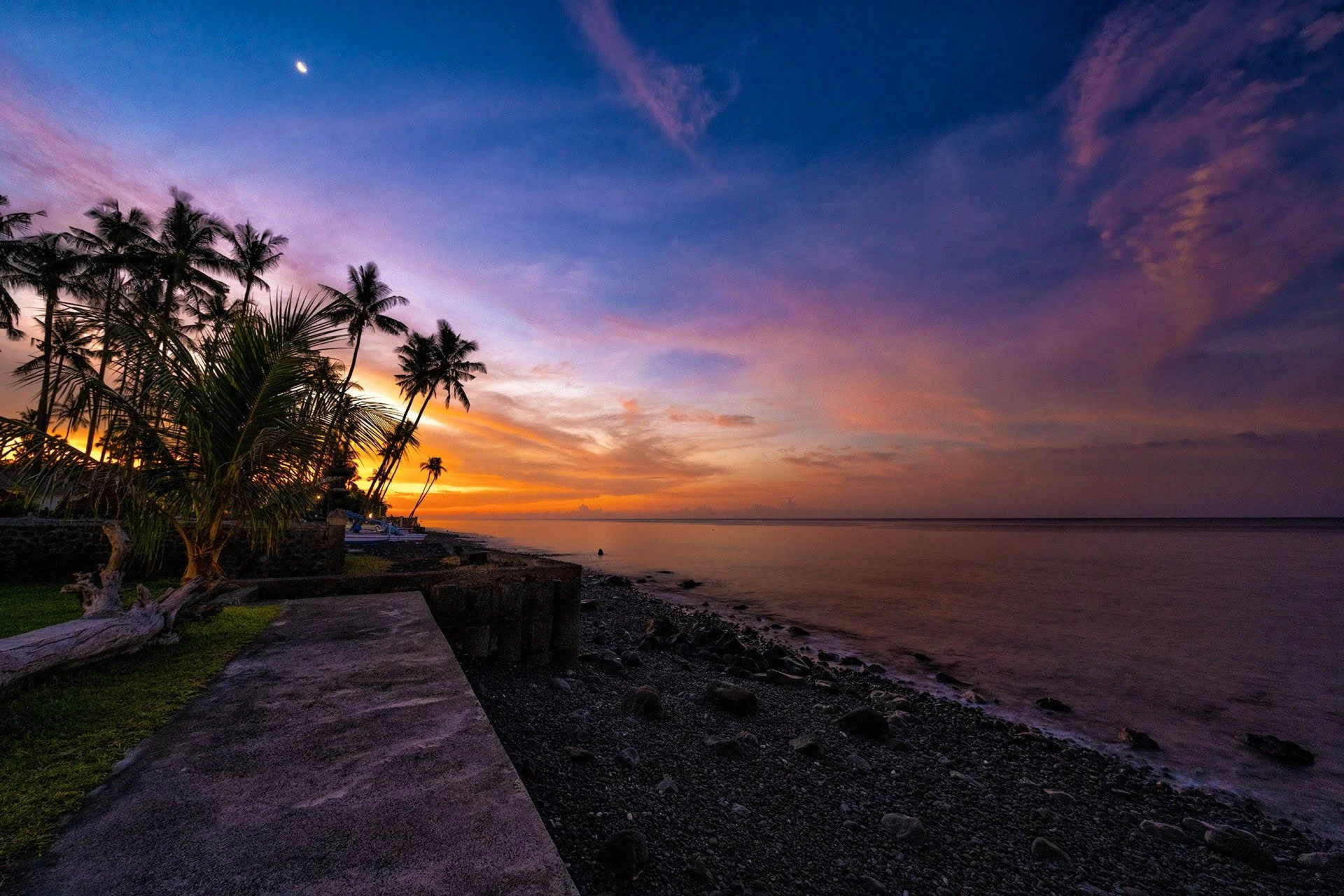 Solnedgang ved kyst på Bali - himlen brydes i mange flotte farverog bølgerne slå stille mod strandkanten der består af små sten og koralrester - på land læner palmerne sig ud over havet