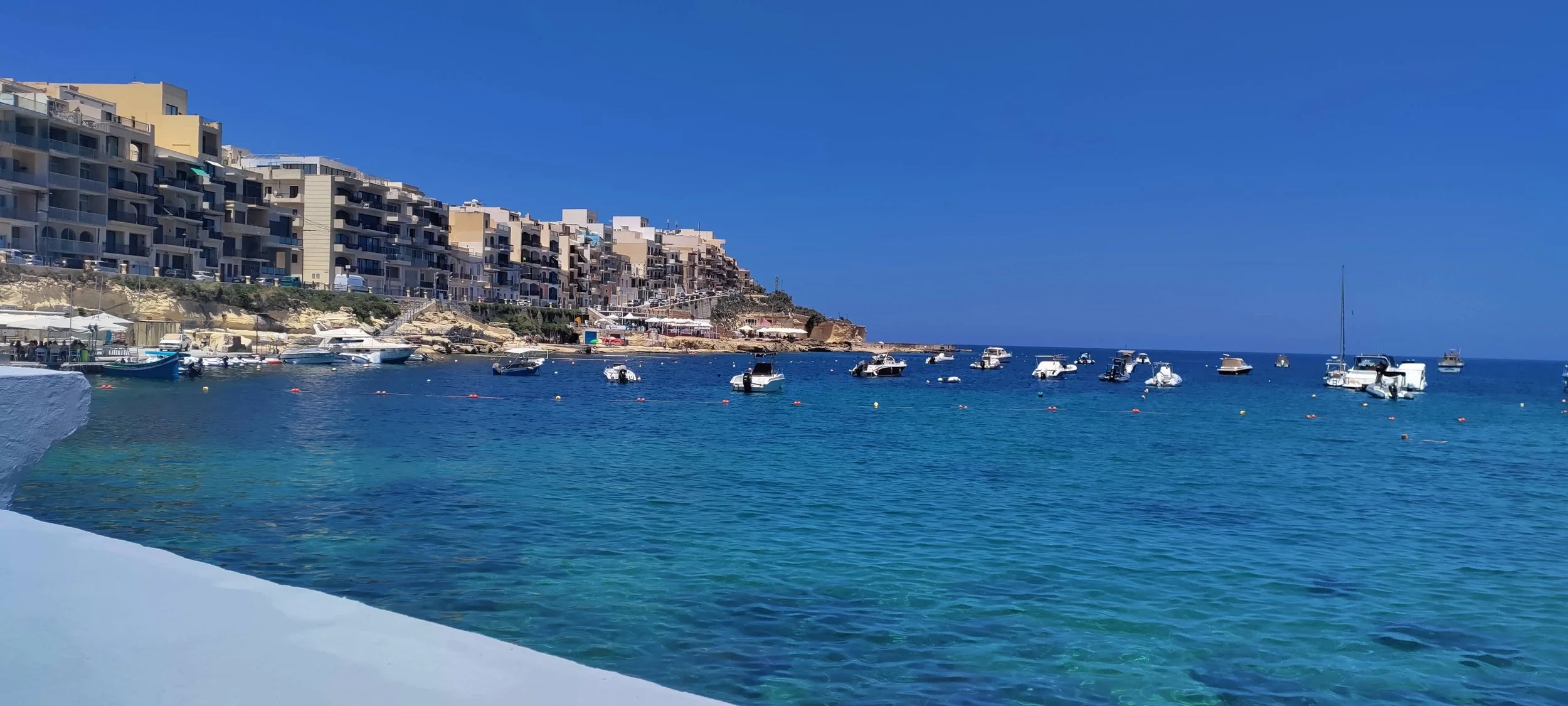 En havn med flere små både og yachts, med en klippe med moderne lejligheder eller hotelbygninger i baggrunden under en klar blå himmel.