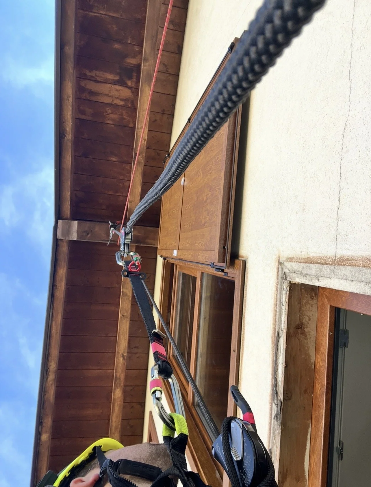 L'immagine mostra una persona con imbracatura di sicurezza, utilizzando un sistema di carrucole per un'attività di lavoro in quota. La scena si svolge sul balcone di un edificio con pareti di cemento, caratterizzato da un tetto di legno.