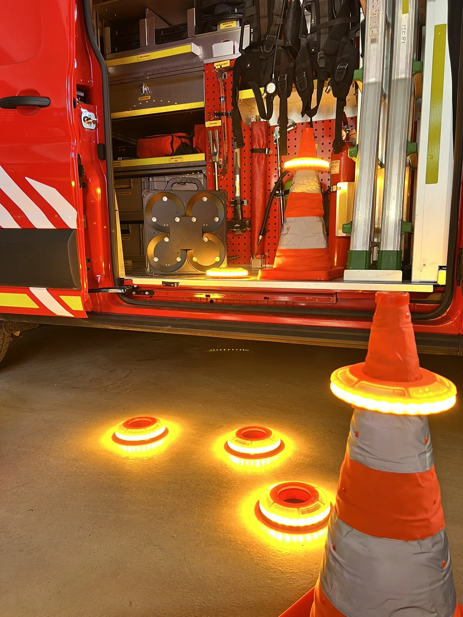 L'interno di un veicolo dei vigili del fuoco con attrezzi e attrezzature di emergenza, tra cui barriere di sicurezza luminose e coni di segnaletica, visibili anche alcuni coni di traffico con luci arancioni accese posizionati sul pavimento.
