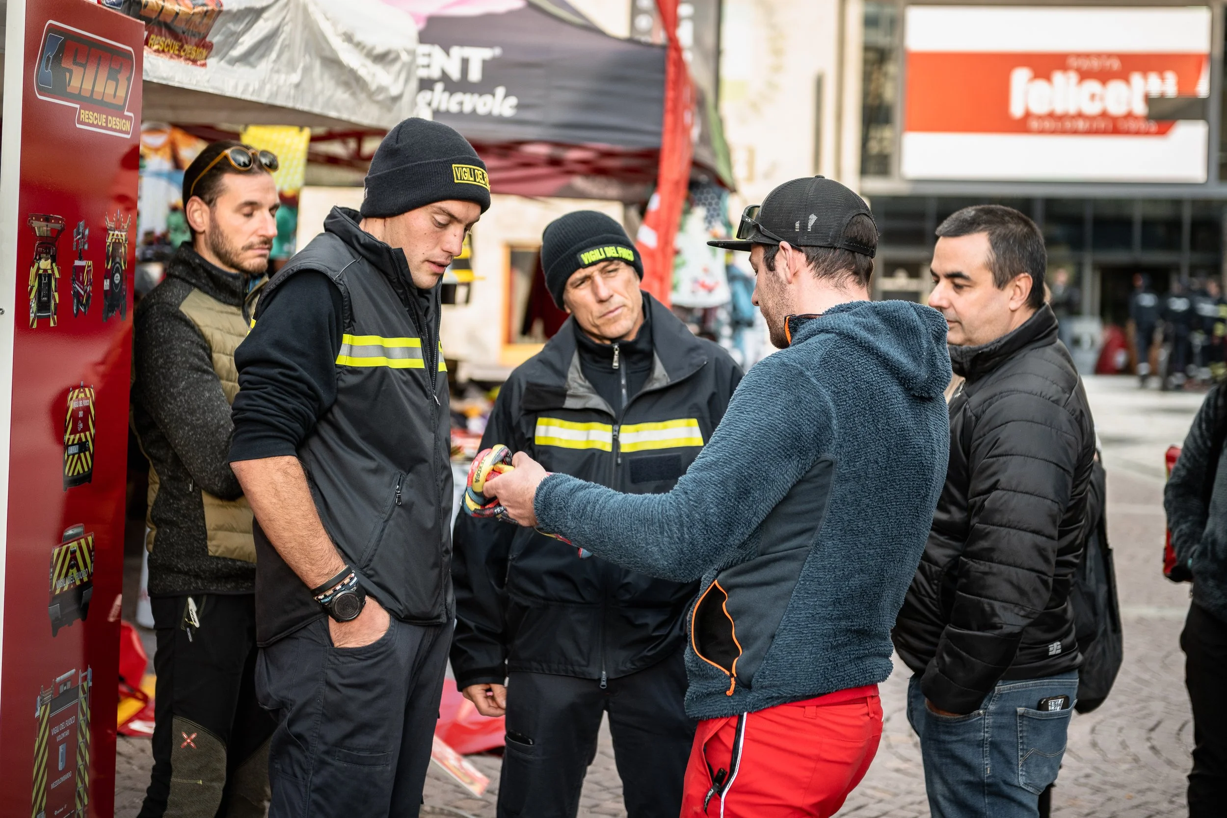 Gruppo di cinque uomini che discutono, alcuni indossano divise da vigili del fuoco e cappelli con scritto "Vigili del Fuoco" in un'area all'aperto con stand e cartelloni pubblicitari.
