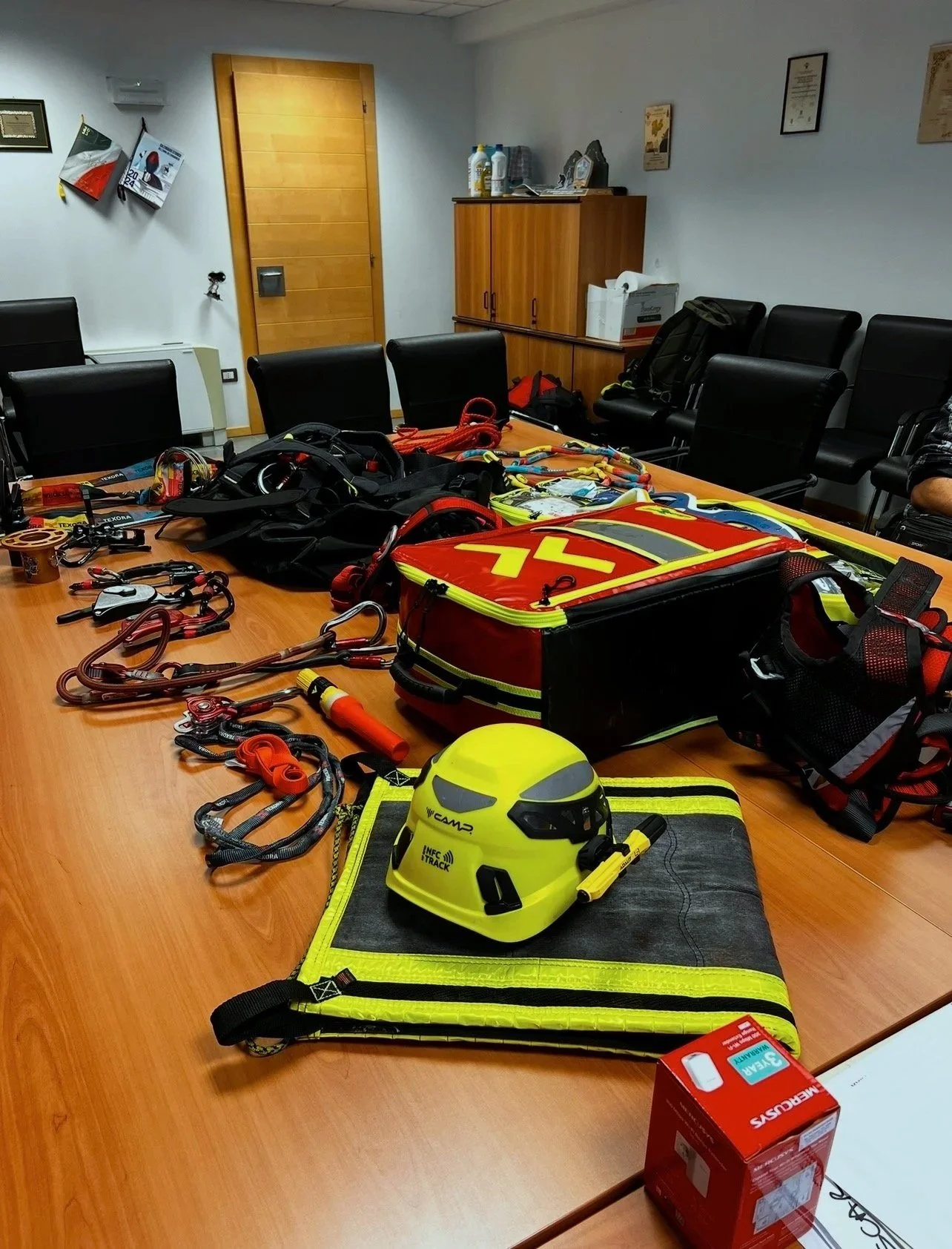Attrezzature di emergenza e sicurezza in un ufficio, tra cui una tuta da elisoccorso, casco di sicurezza, corde, imbragature e altri strumenti di soccorso.