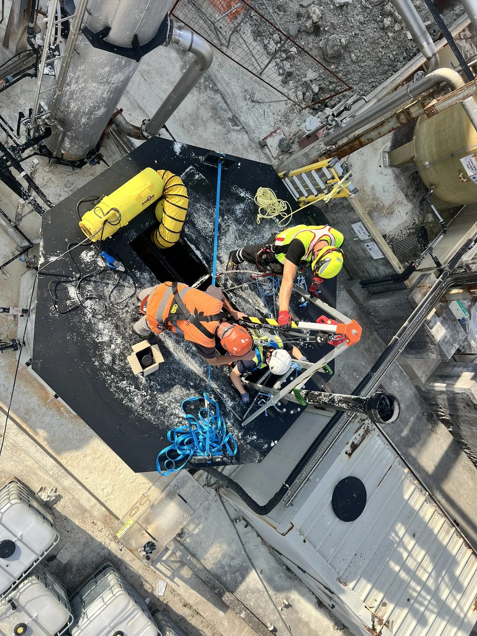 Operai al lavoro su una piattaforma di manutenzione in un cantiere industriale, indossano caschi e dispositivi di protezione, con attrezzature e tubi metallici visibili nell'ambiente circostante.