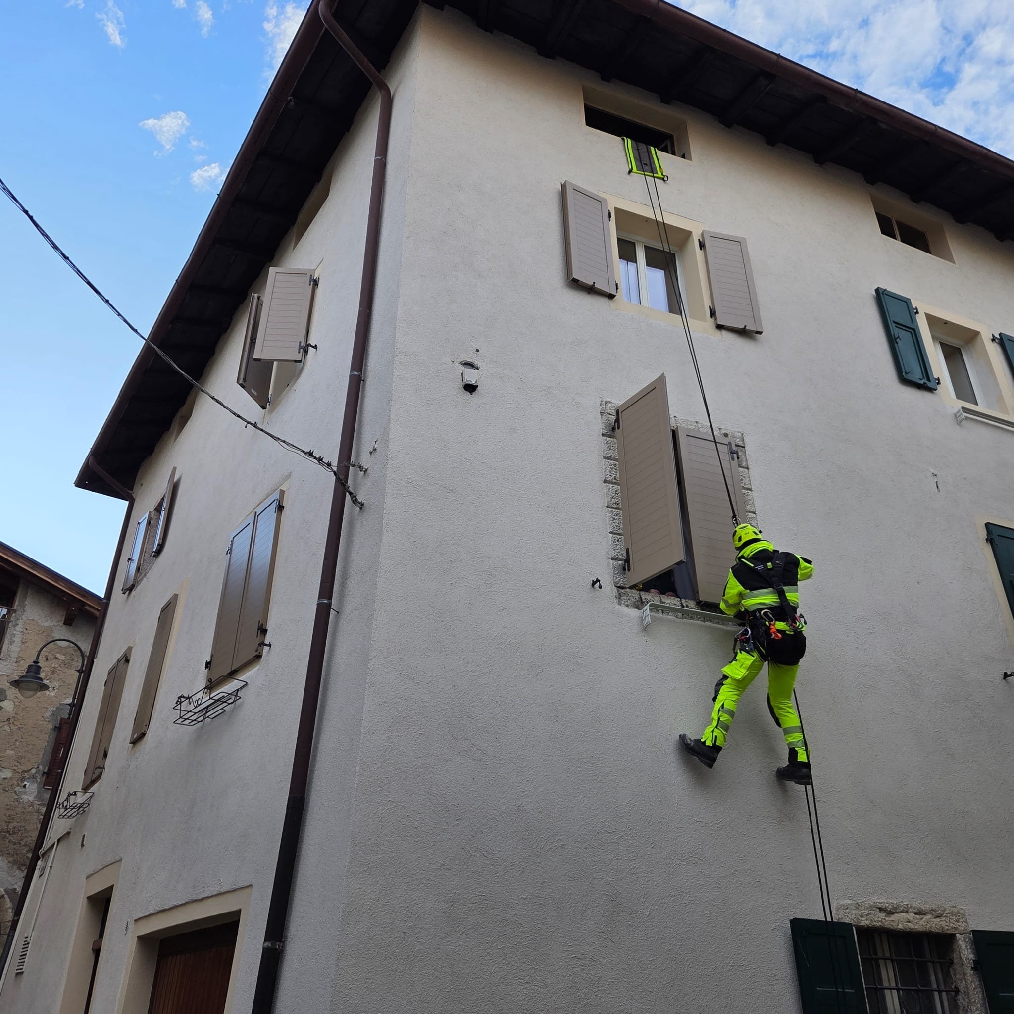 Un soccorritore vestito di giallo con equipaggiamento si arrampica su una corda che scende da una finestra di un edificio di multi piani. La scena si svolge di giorno, con un cielo parzialmente nuvoloso.
