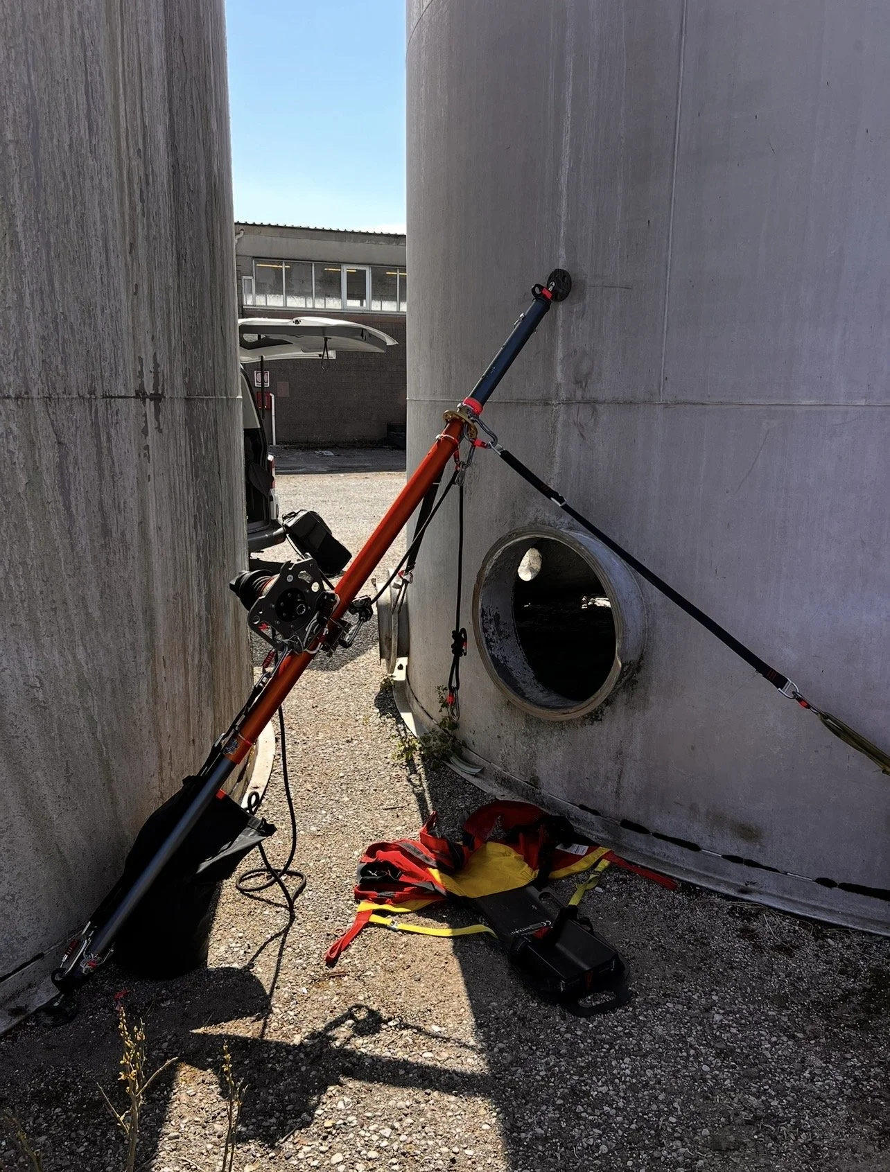 Una fotocamera, un dispositivo elettronico e un bastone lungo e colorato appoggiati tra due grandi pareti di cemento, con alcuni strumenti e una giacca gialla e rossa a terra.