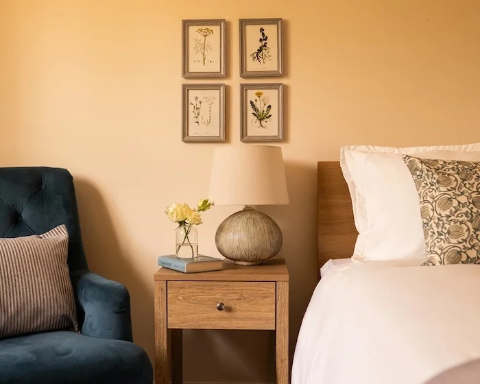 A cozy bedroom corner showing a wooden nightstand with a table lamp, a vase with yellow flowers, and a book. To the left, there is a blue armchair with a pillow. To the right, part of a bed with white bedding and a decorative pillow. Framed botanical art prints are hung on the wall above the nightstand.