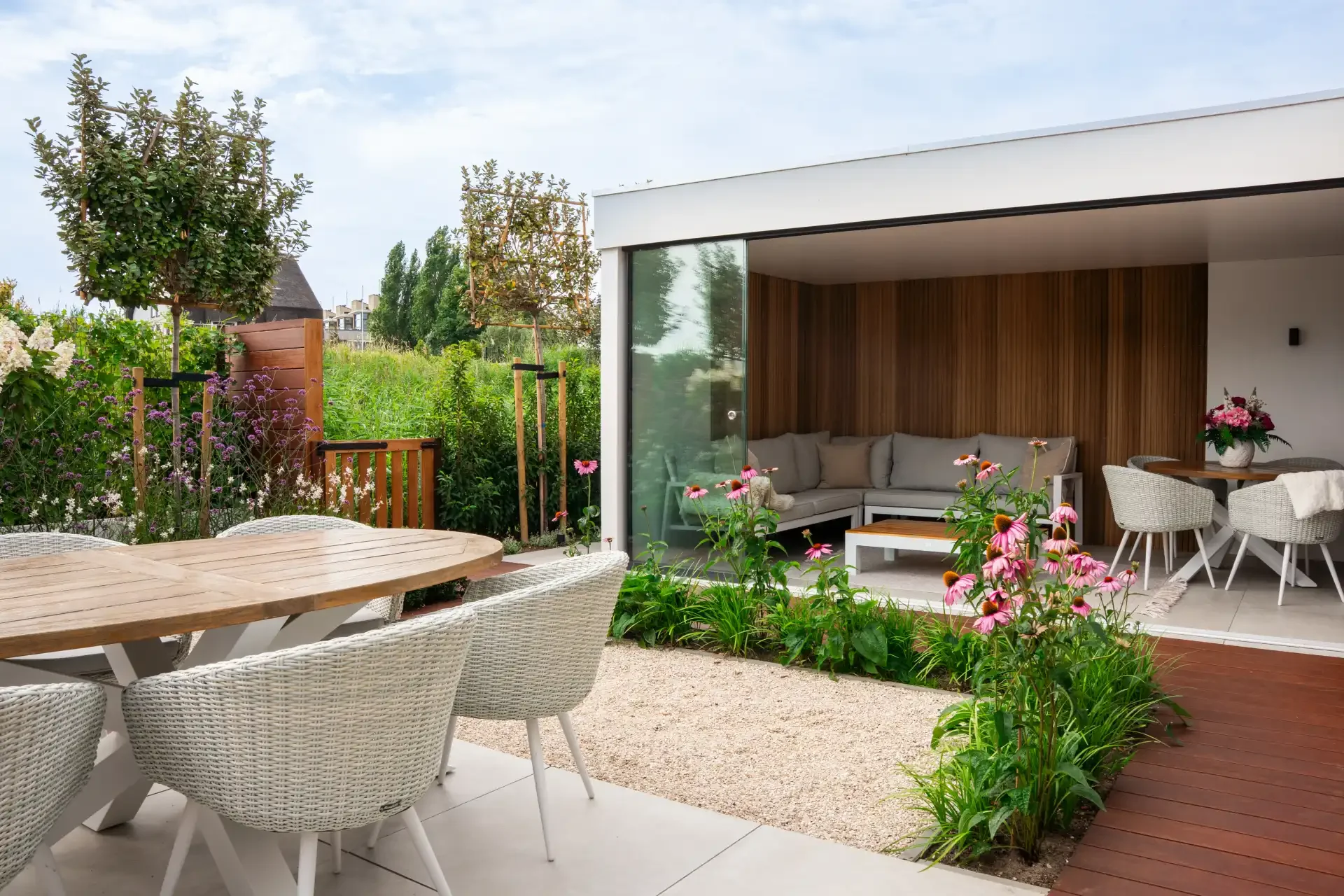 Moderne tuin met houten terras, ronde witte tafel, wicker stoelen, bloemen, tuin en een overdekte zithoek in huis.