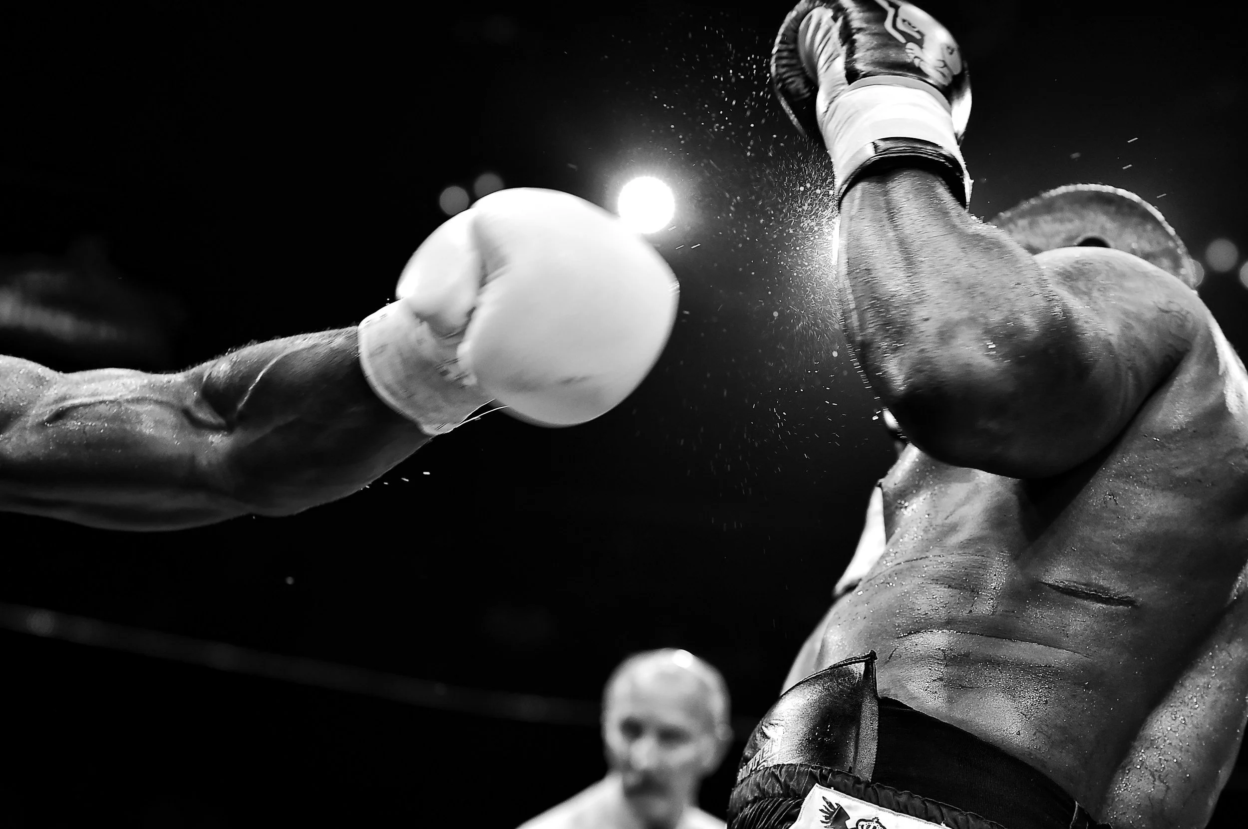 A boxing match scene in black and white, showing a boxer’s upper body and gloves in mid-punch with spray of sweat, with a referee in the background.