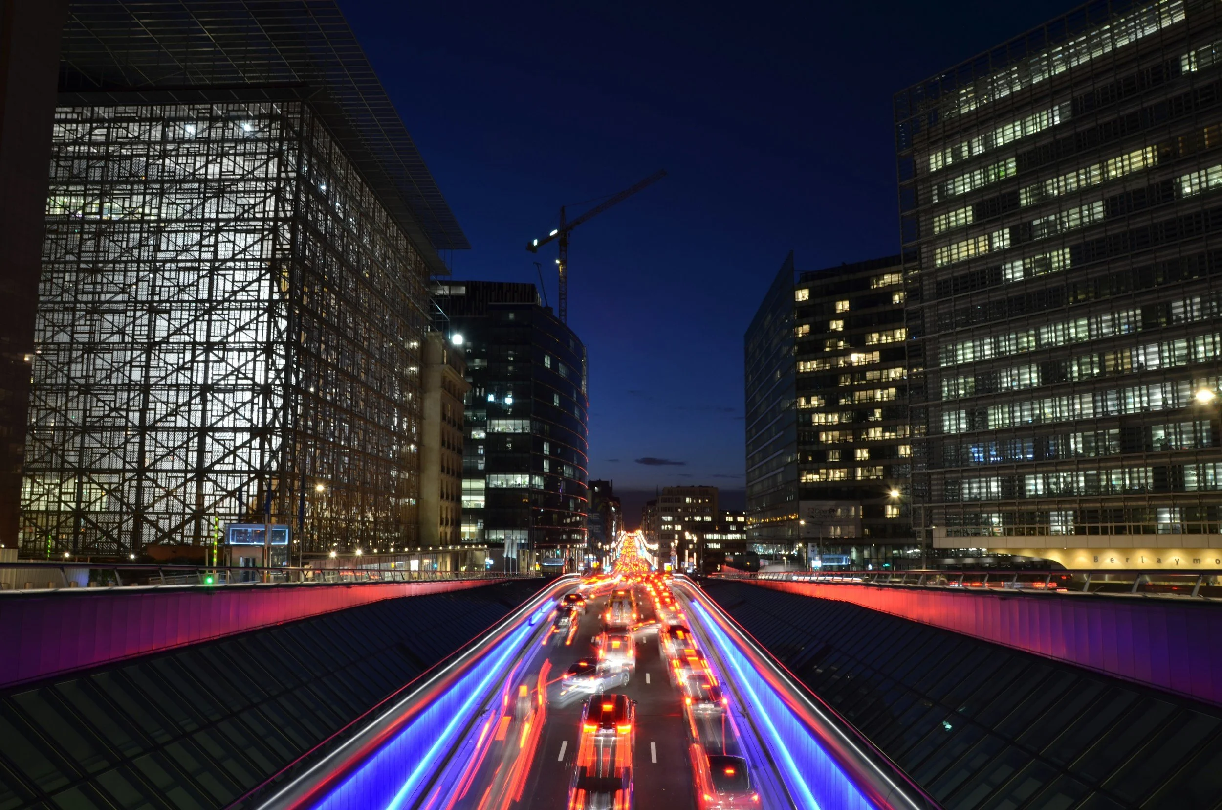 Nighttime cityscape with illuminated buildings, a construction crane, and streaks of red and blue lights from moving cars on a highway