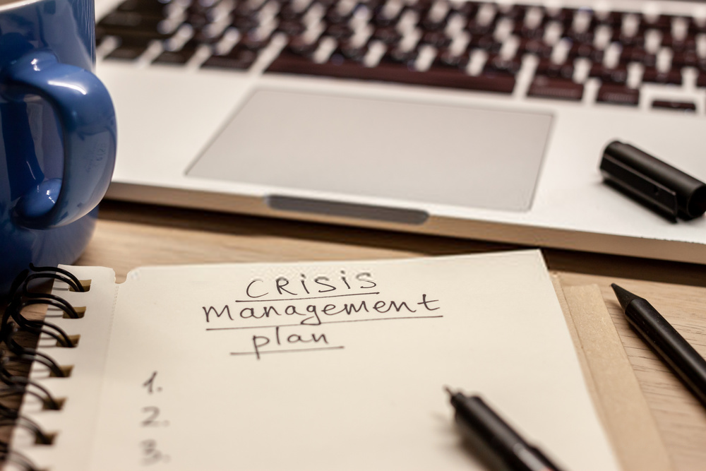 A handwritten crisis management plan on a spiral notebook, with a laptop, mug, pens, and a highlighter on a desk.