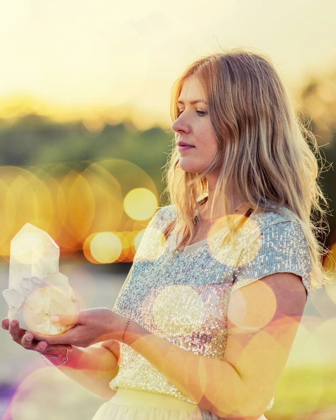 A young woman with wavy blonde hair looking at a small glowing object in her hands during sunset with a blurred background and warm bokeh lights.