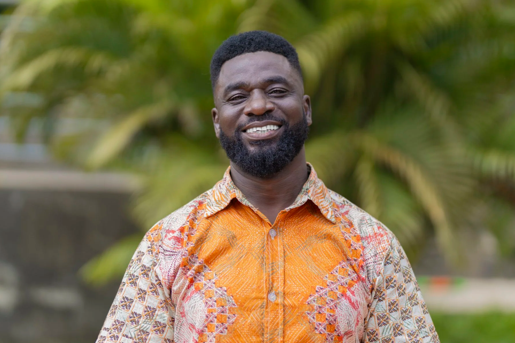 A smiling man with a beard and short hair wearing a colorful patterned orange and white shirt, standing outdoors with green foliage in the background.