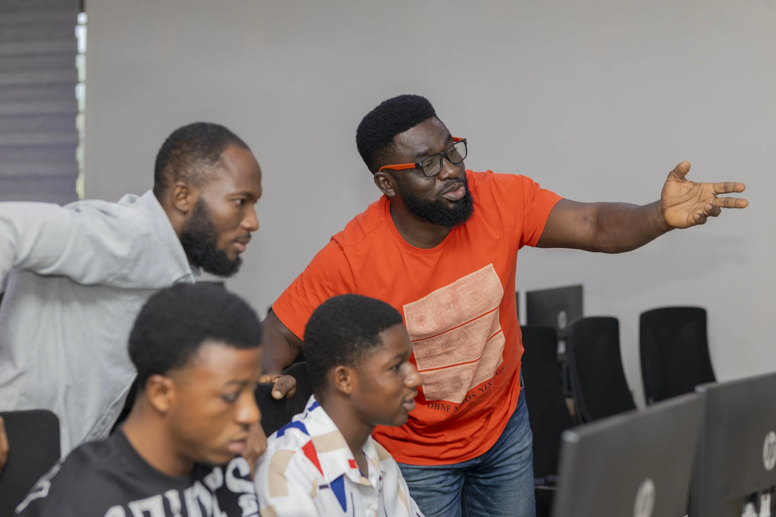 A man with glasses and an orange shirt is giving instructions or explaining something to three young men in a room with computers.