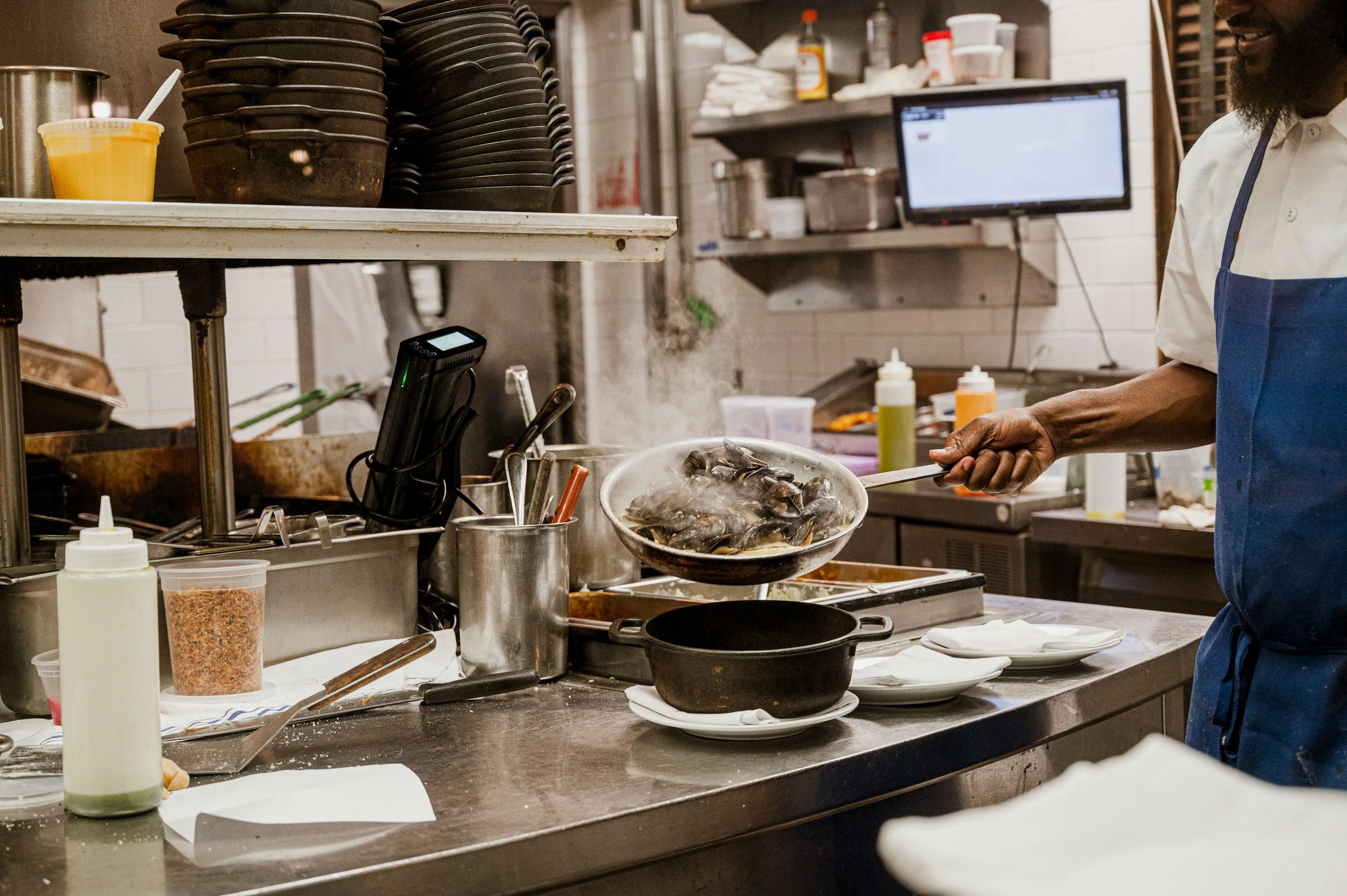 A chef cooking mussels in a kitchen, using a frying pan over a stove, with various kitchen tools and ingredients around.