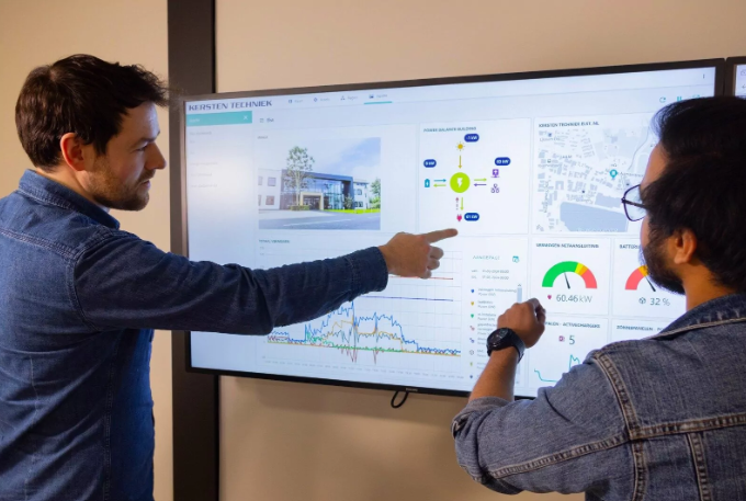 Two men discussing data and graphs displayed on a large screen in an office setting.