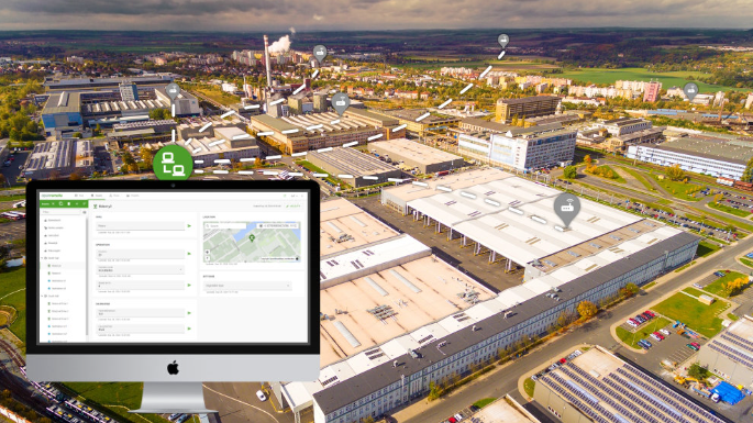 Aerial view of a large industrial complex with many warehouse buildings, parking lots, and roads. Overlaid is a computer monitor showing a logistics or management software interface with a map. The scene is set outdoors with a cloudy sky and distant landscape.