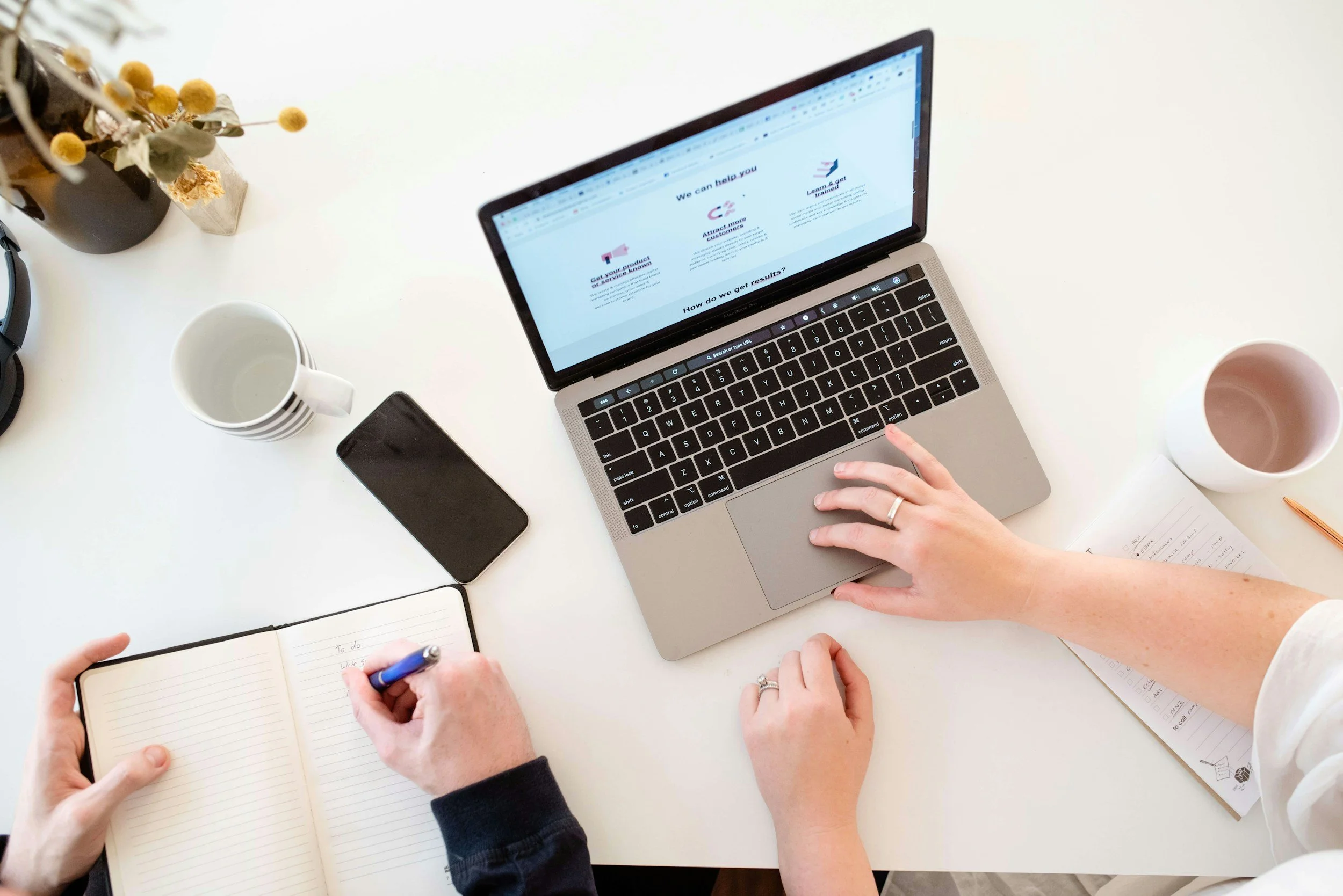 Overhead view of a desk with two people working. One person is writing in a notebook with a pen, and the other is using a laptop. The desk has coffee mugs, a smartphone, a notebook, a pen, a paper, and a vase with dried flowers.