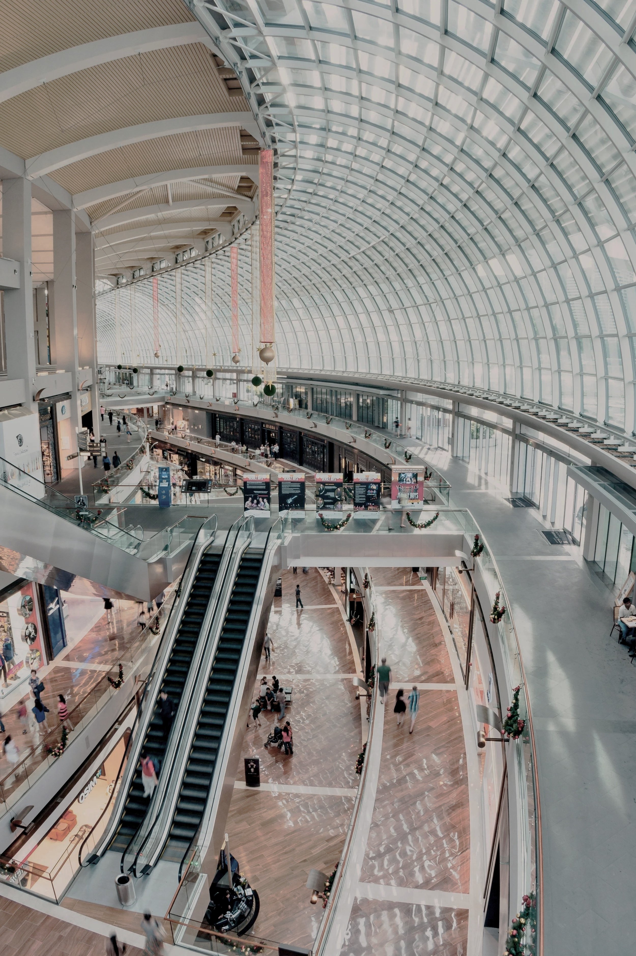 Interior view of a modern shopping mall with a large curved glass ceiling, escalators, shoppers, and Christmas decorations.