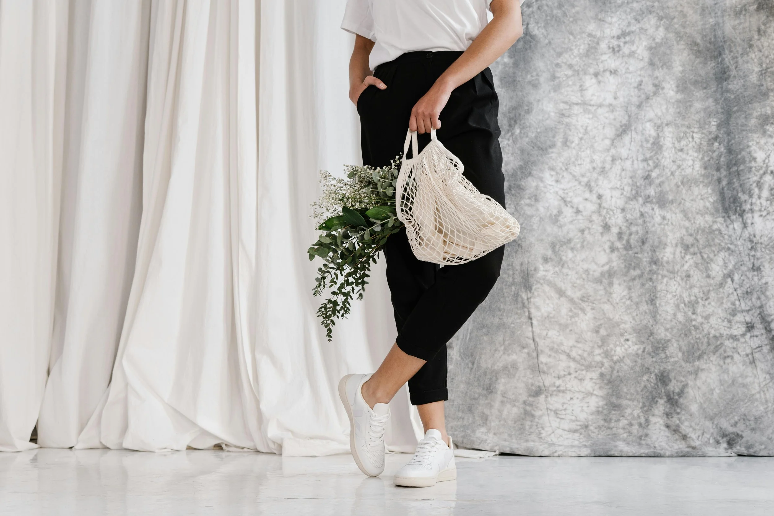 Person standing with one hand in pocket, holding a mesh tote bag with greenery, dressed in white t-shirt, black pants, and white sneakers, standing in front of a curtain and textured gray background.