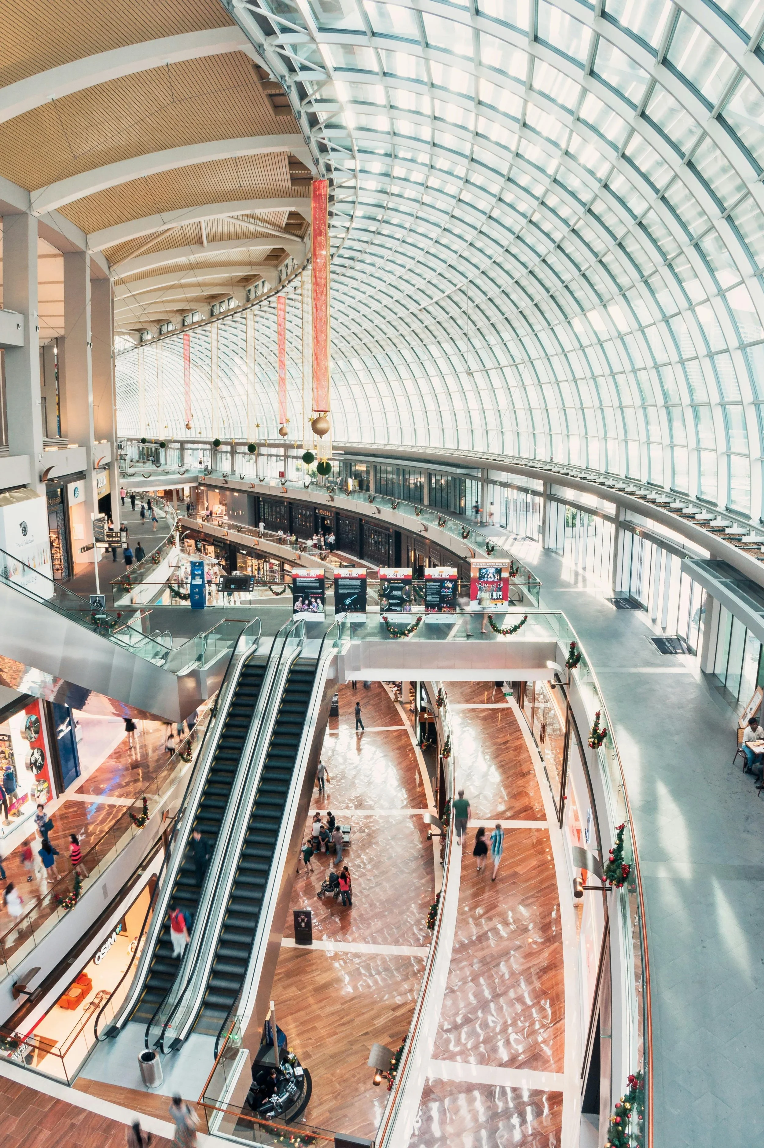 Inside view of a modern shopping mall with large arched glass ceiling, multiple floors, escalators, stores, and shoppers.