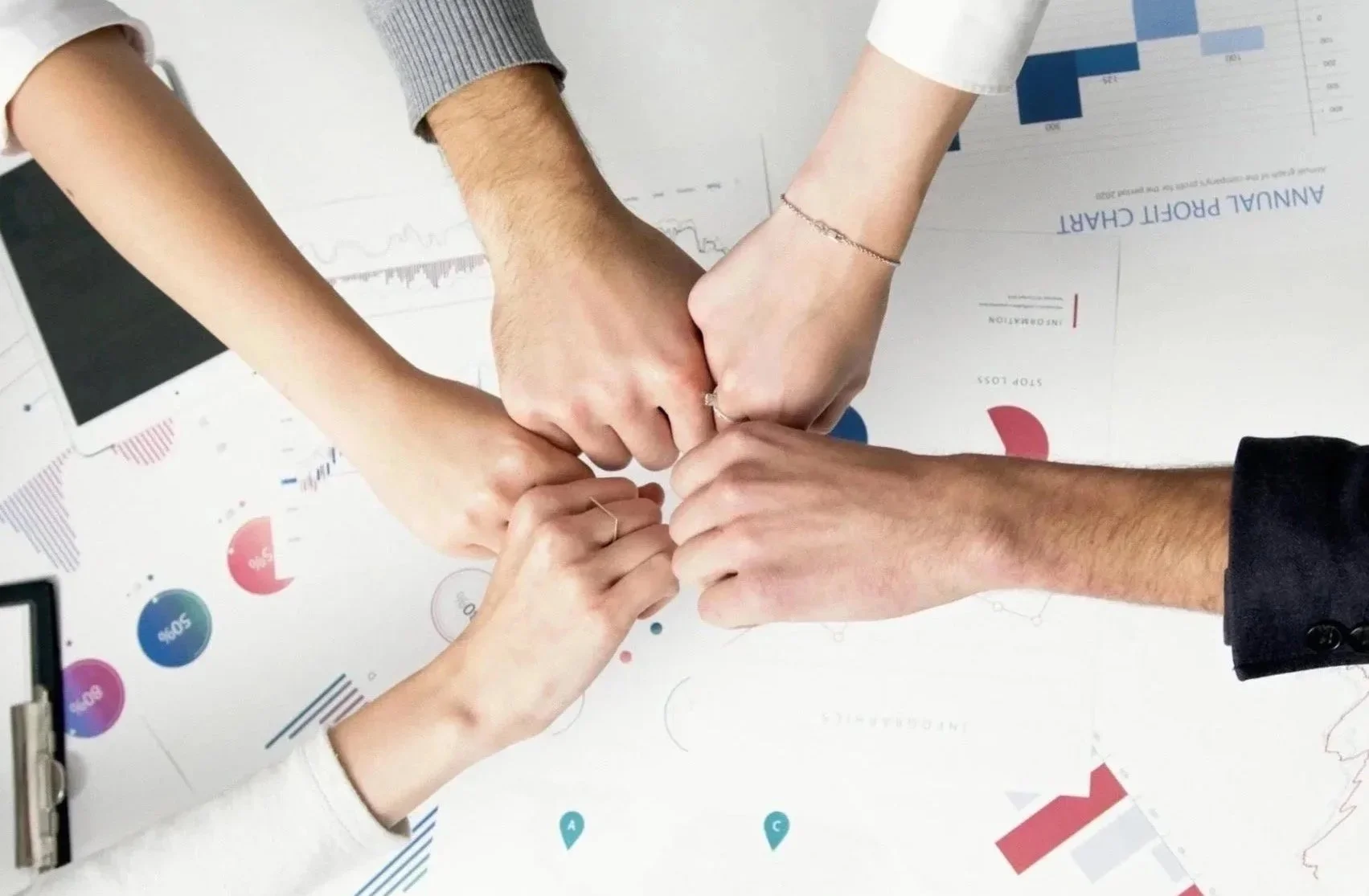Four people with different skin tones join their hands together in a show of teamwork, over a table with charts and a tablet.