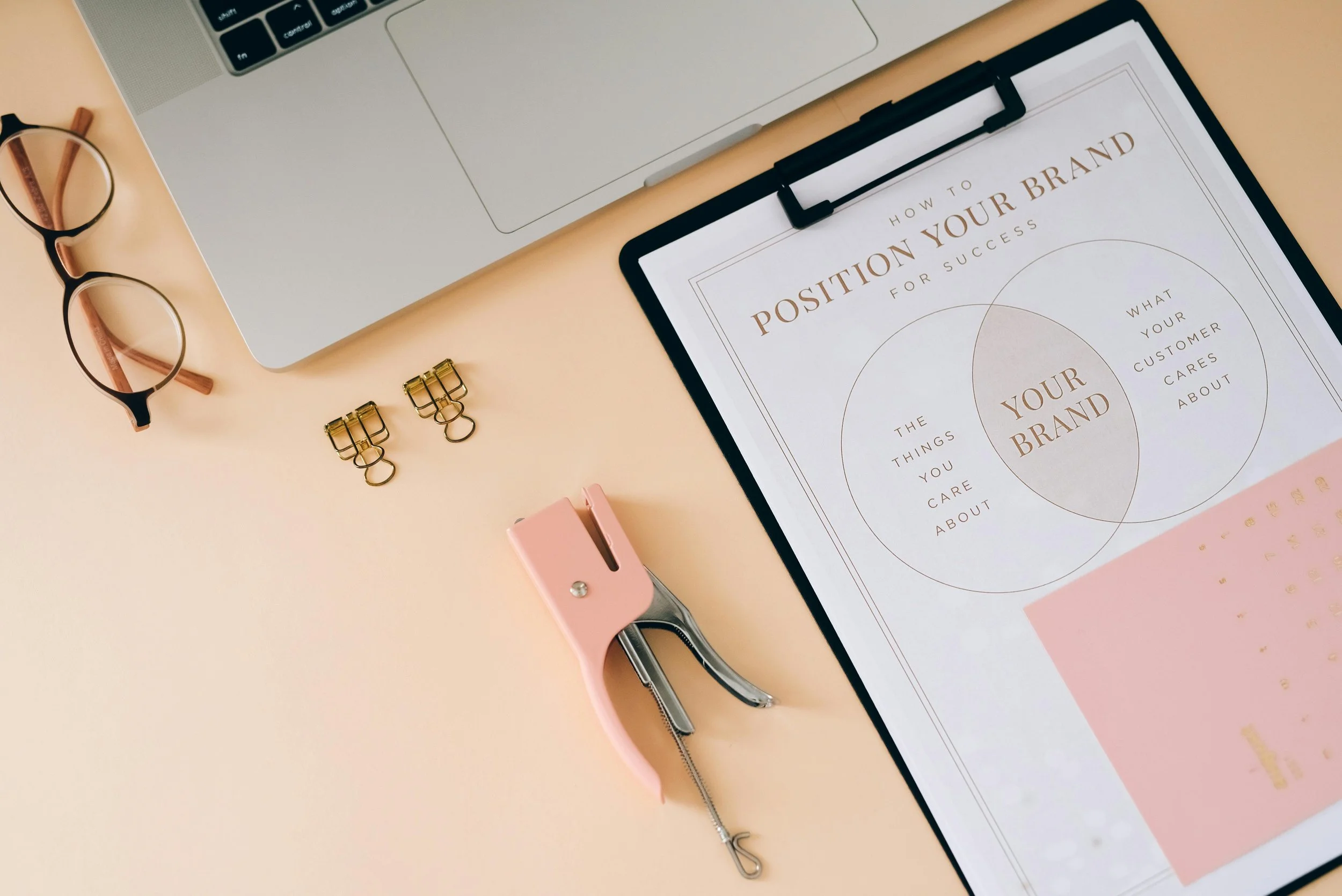 A flat lay of a workspace including a silver laptop, a pair of black-rimmed glasses, a pink stapler, three gold binder clips, a clipboard with a paper titled "How to Position Your Brand for Success," a pink pen, and a pair of scissors on a light peach-colored surface.