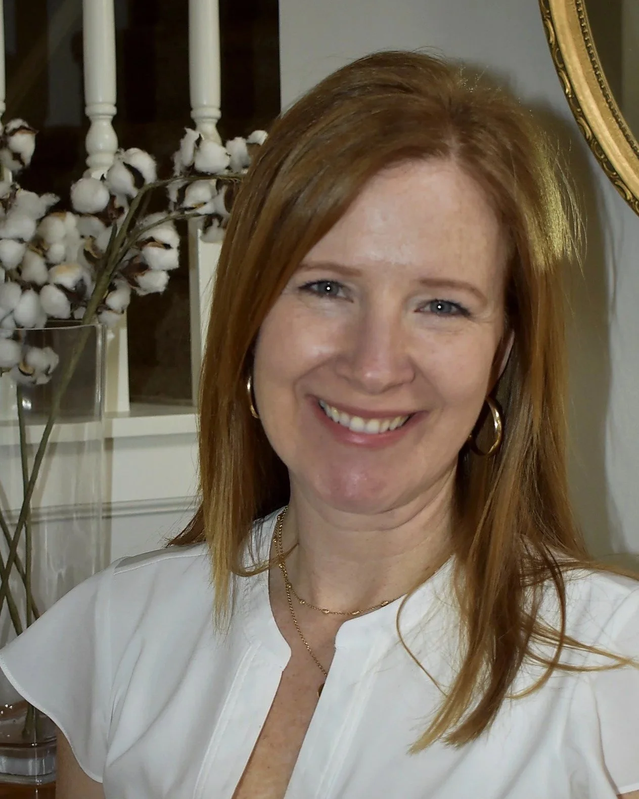 A woman with shoulder-length red hair smiling at the camera inside a house with decorative cotton plant arrangements and a mirror in the background.