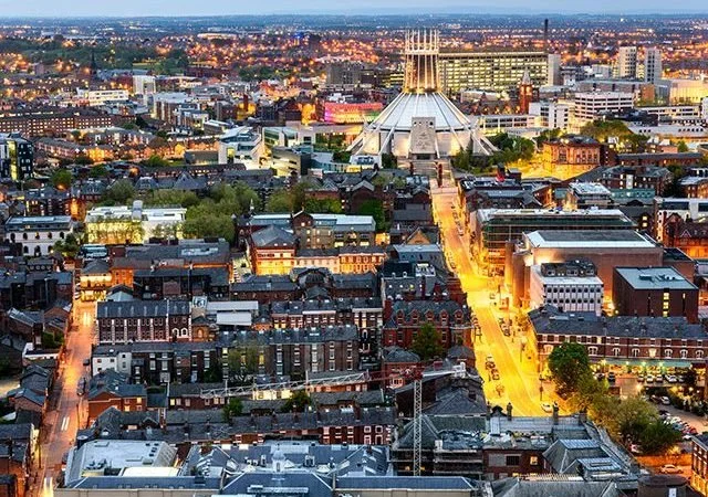 Aerial view of a cityscape at dusk with a mix of old and modern buildings, illuminated streets, and a prominent white, tent-like structure in the distance.